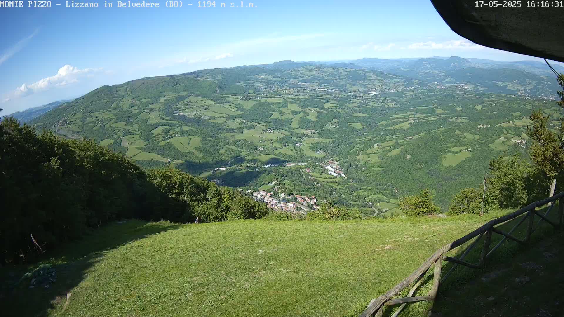 A panoramic view shows a verdant valley dotted with small towns and farmlands under a mostly clear blue sky.