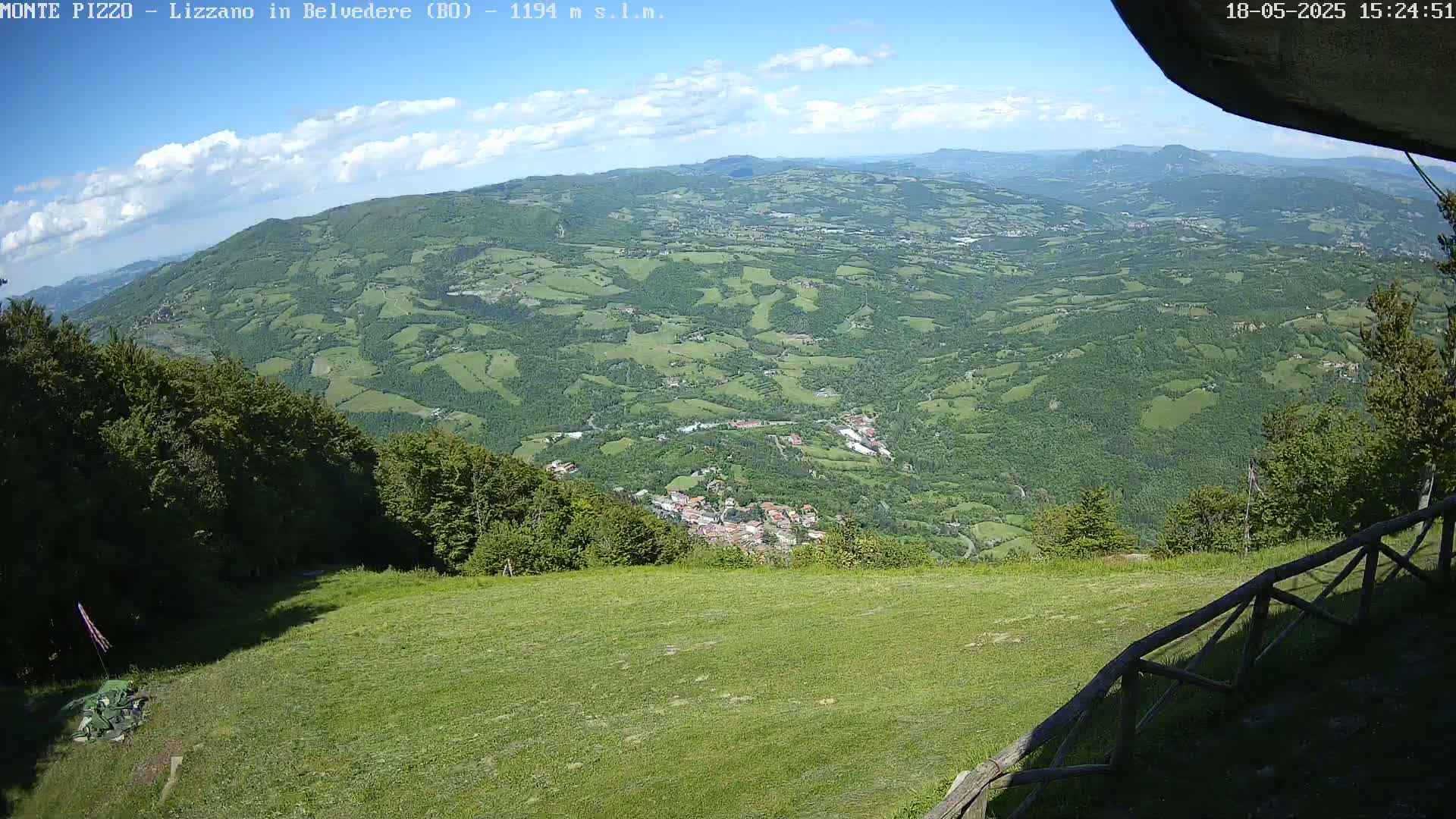 A sprawling green valley dotted with small towns and farmland stretches out beneath a partly cloudy blue sky, viewed from a grassy hilltop with a wooden fence.
