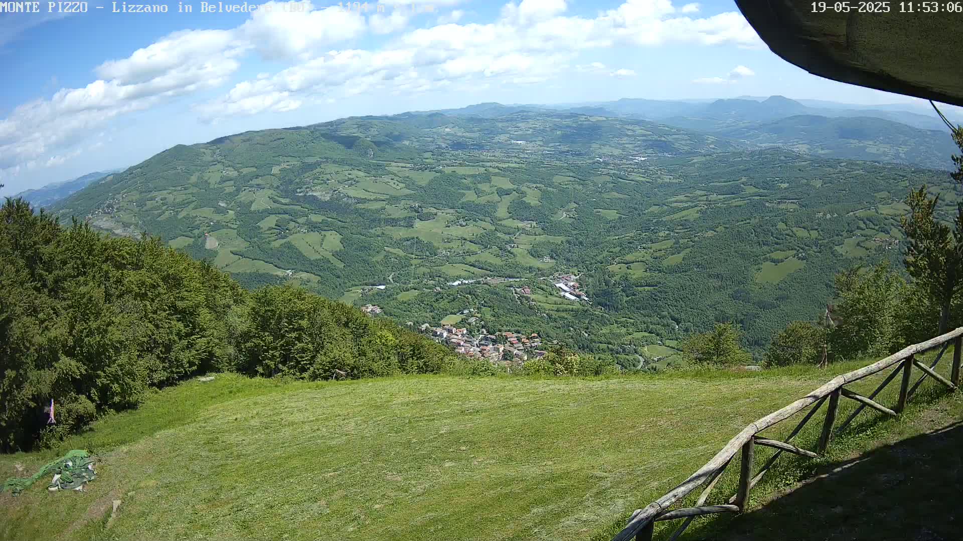 A partly cloudy sky overlooks a vast green valley, containing a small town nestled amongst the hillsides, from a grassy hilltop with a wooden fence.