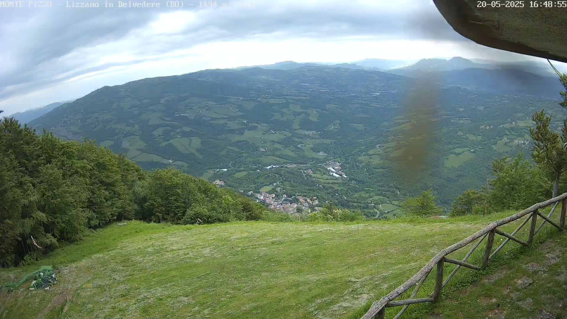 A grassy hilltop overlooking a valley of green hills and a small town under a partly cloudy sky.