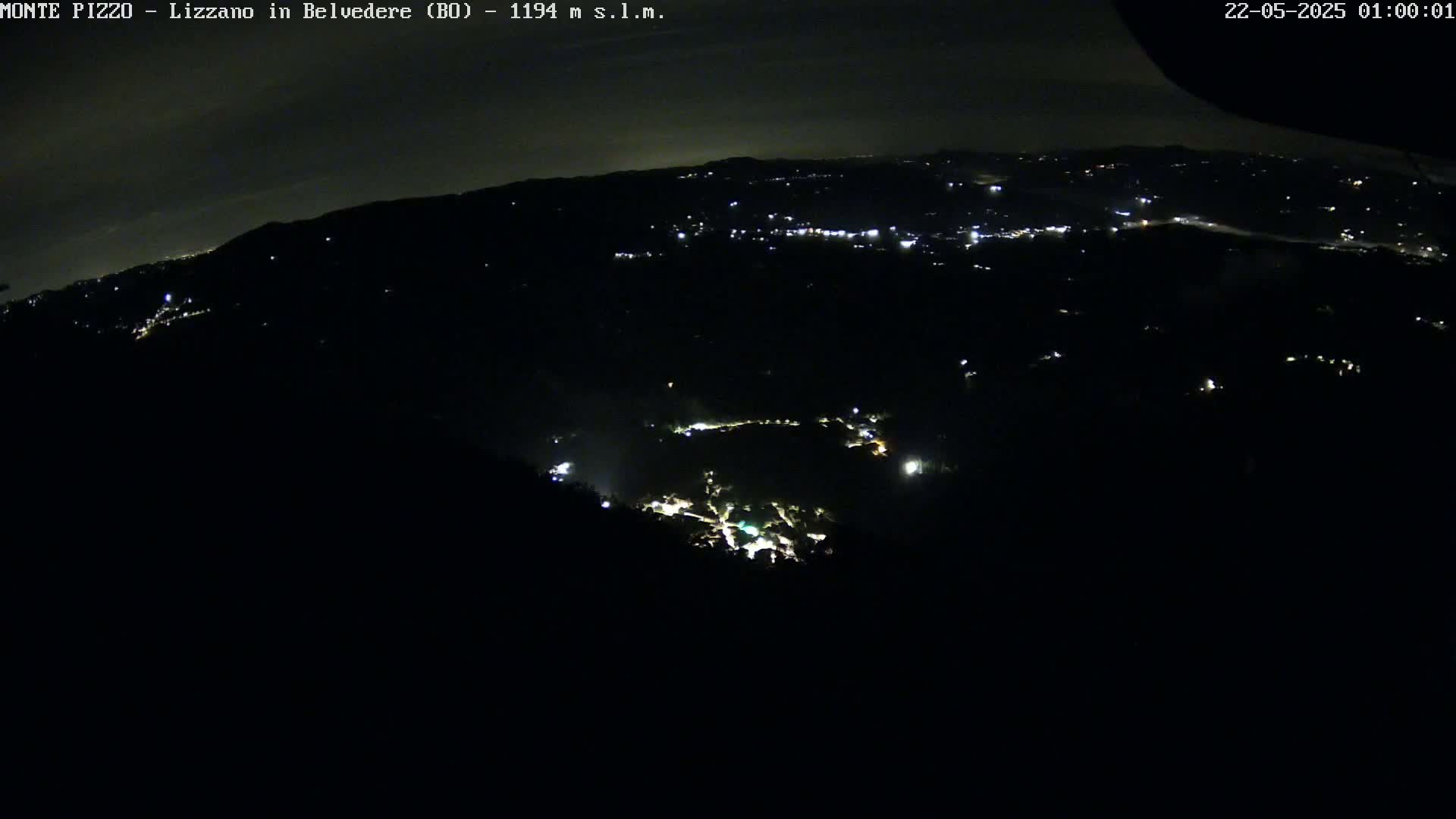 A nighttime, high-angle view shows a dark, hilly landscape sparsely dotted with faint lights from a distant town.