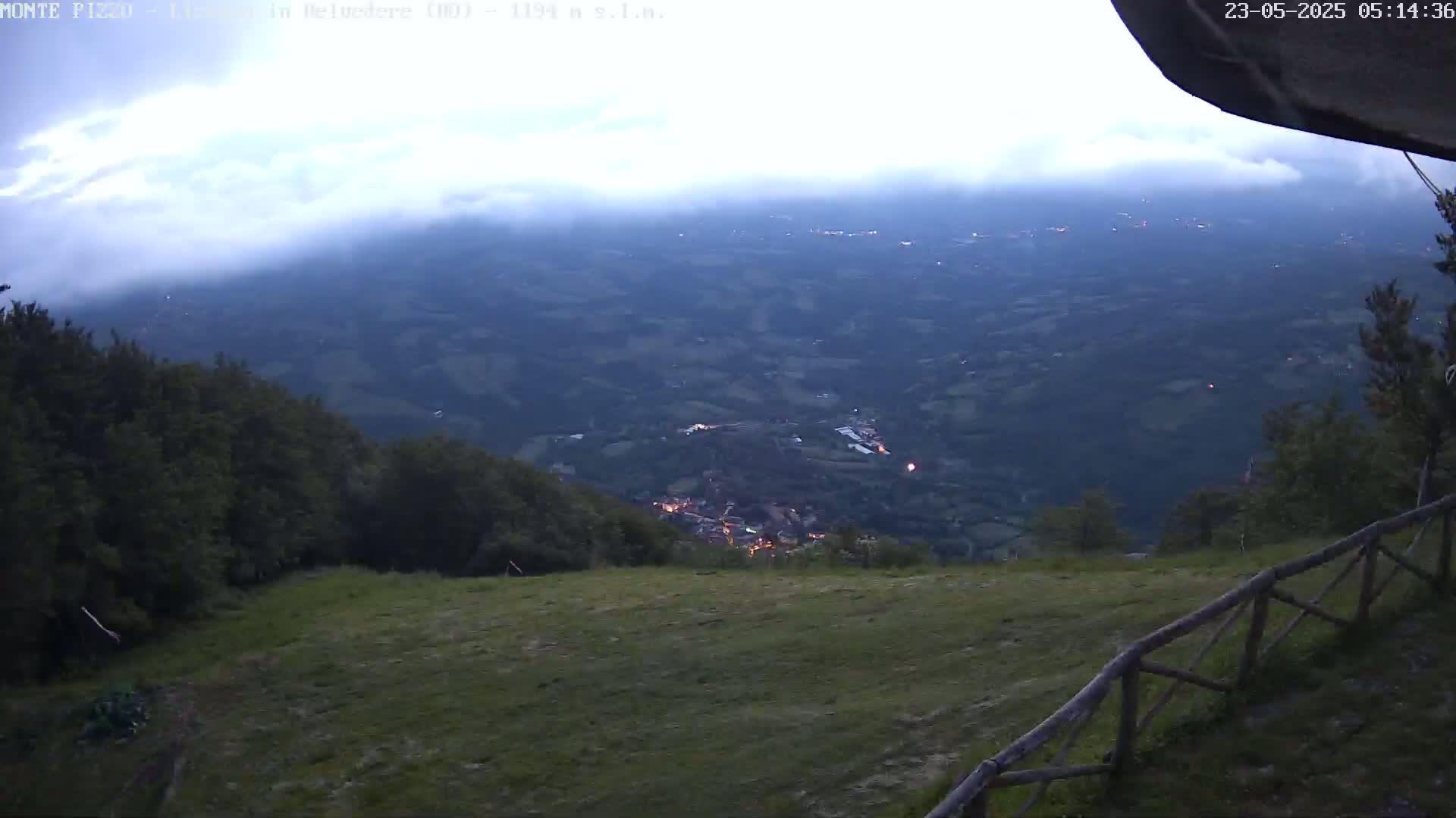 A high-angle, twilight view from a mountaintop shows a valley below with a small village illuminated at dusk, partially obscured by fog or clouds.