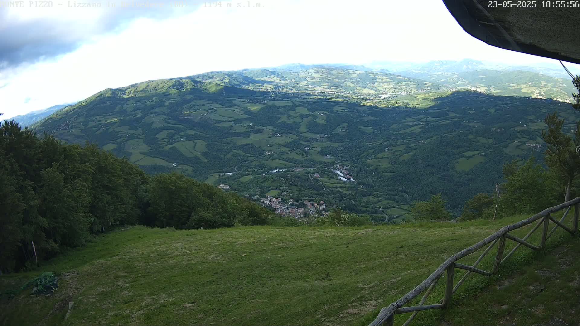 A sprawling green valley dotted with small towns and villages is viewed from a grassy hilltop under partly cloudy skies.