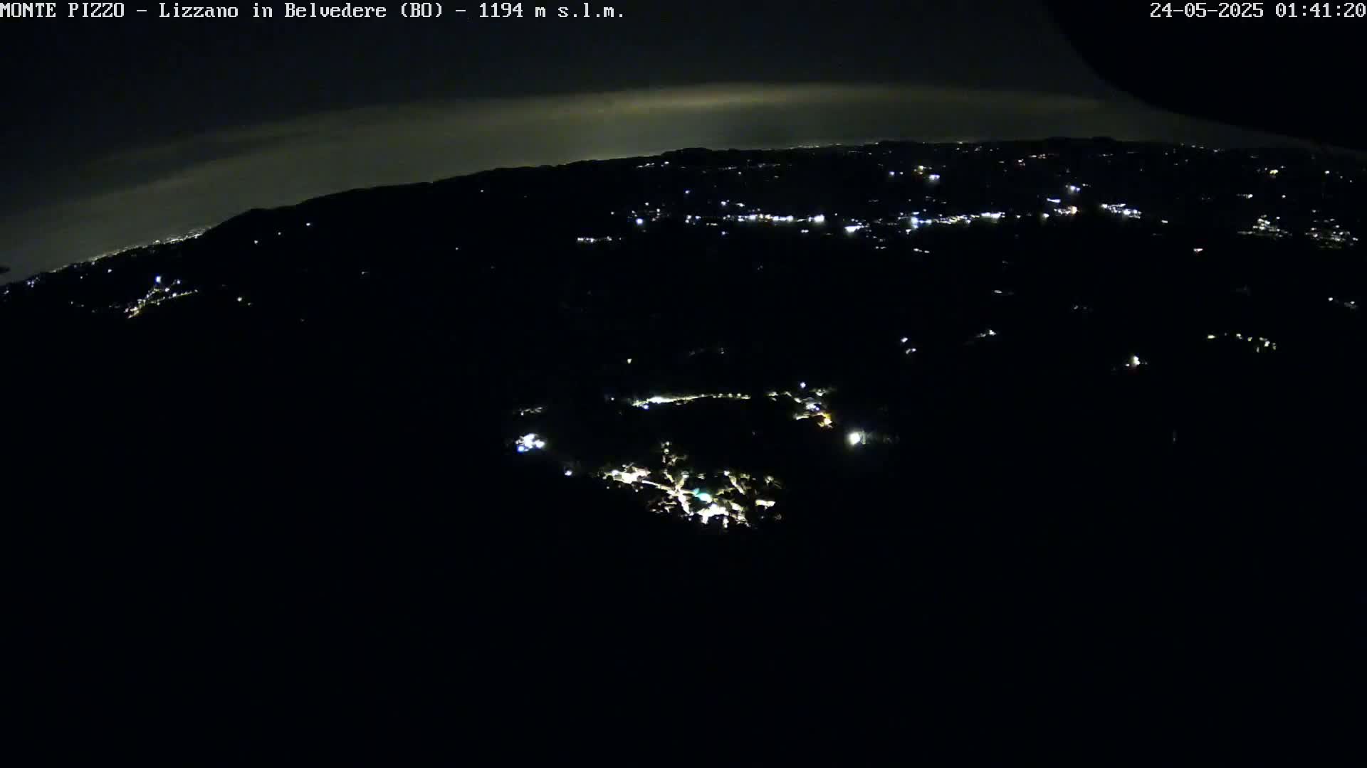 A nighttime, high-angle view shows a dark, hilly landscape sparsely dotted with small, scattered lights, extending to a distant horizon illuminated by a faint glow.