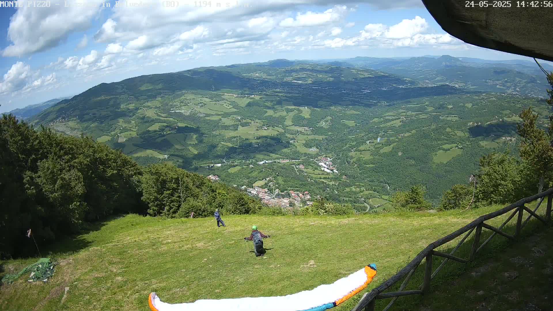 Two people prepare to paraglide on a grassy hilltop overlooking a vast, verdant valley under a partly cloudy sky.