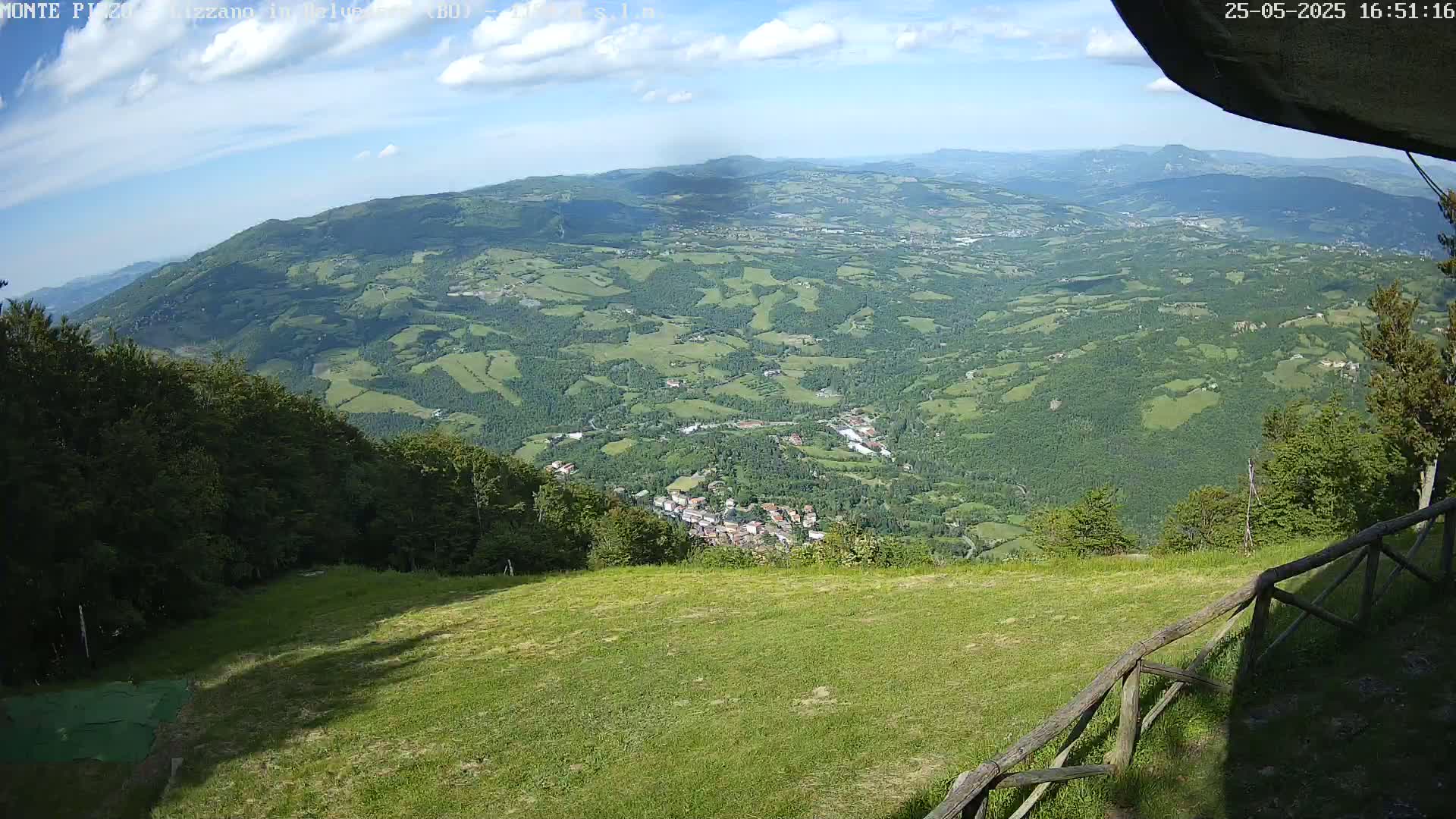 A grassy hilltop overlooking a valley of rolling green hills and a small town under a partly cloudy sky.