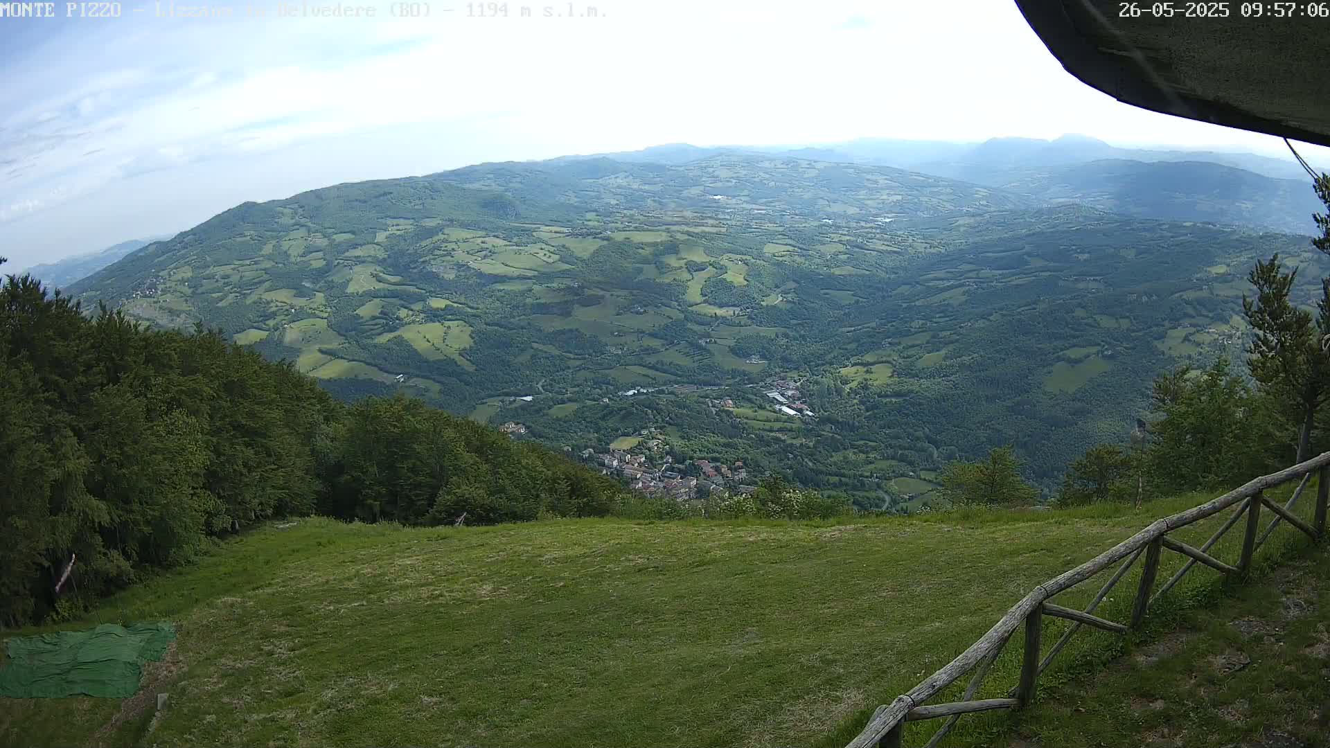 A sprawling green valley dotted with small towns and villages stretches out beneath a mostly clear sky, viewed from a grassy hillside with a wooden fence.