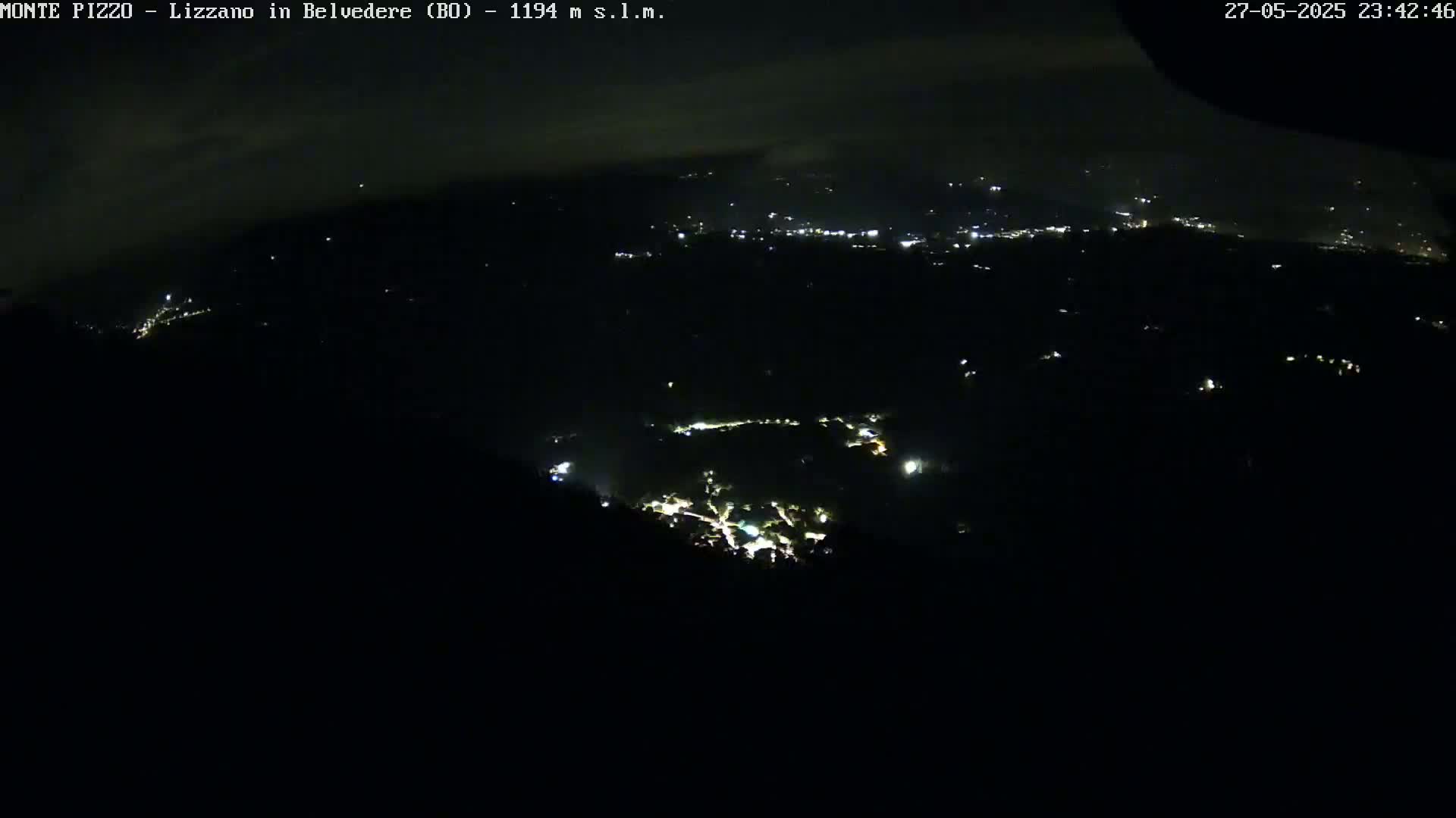 A nighttime, high-angle view shows a valley with scattered lights, under a slightly cloudy sky.