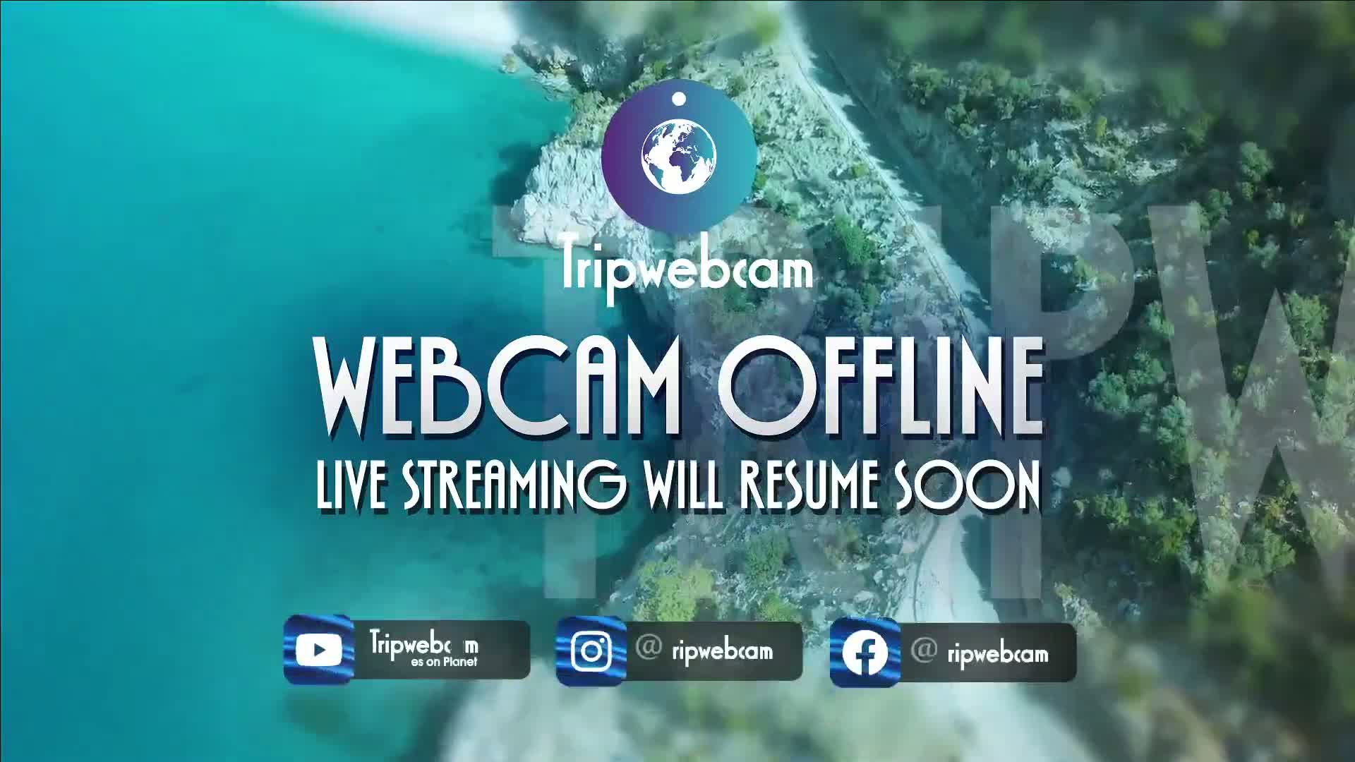 An aerial view shows a tranquil coastline with turquoise water and lush green vegetation, overlaid with a message indicating the webcam is temporarily offline.