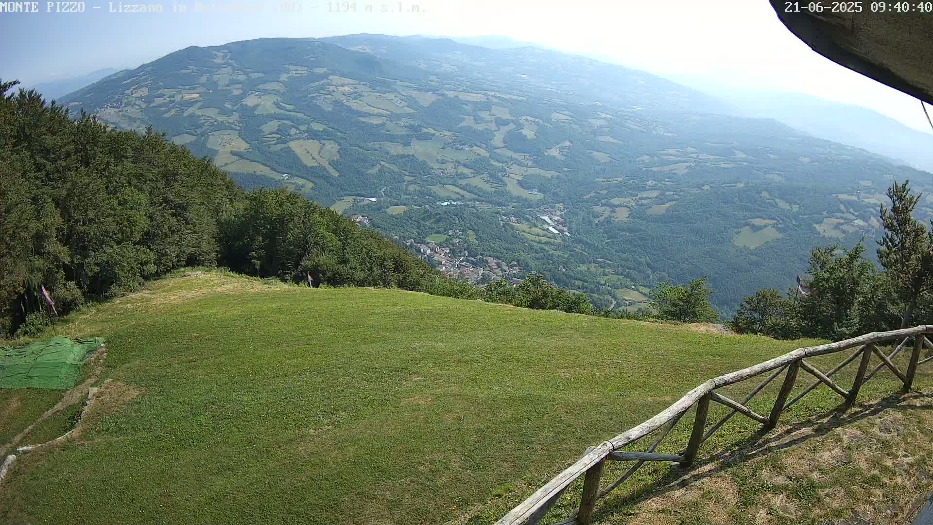 A grassy hilltop with a wooden fence overlooks a valley of rolling hills and farmland under a clear sky.