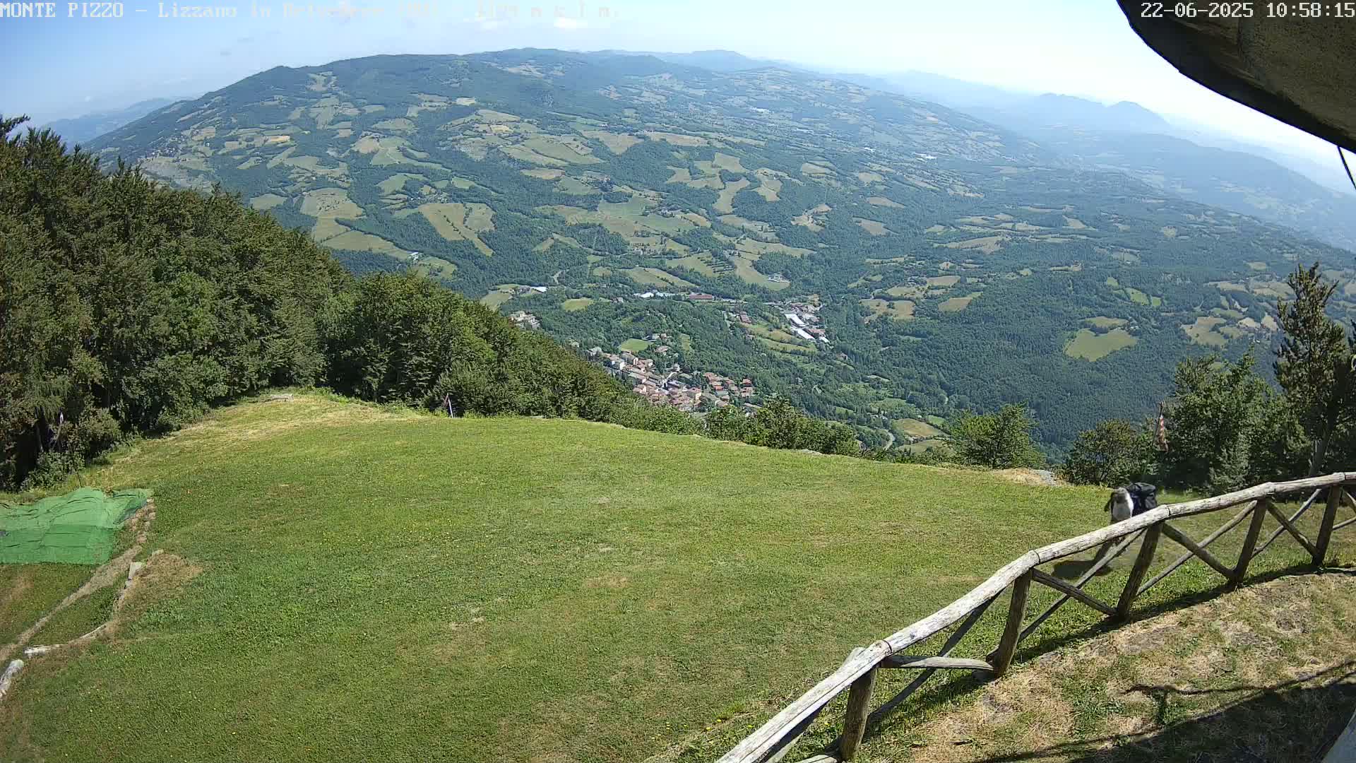 A grassy area with a wooden fence overlooks a vast, sunny valley dotted with trees and small settlements.