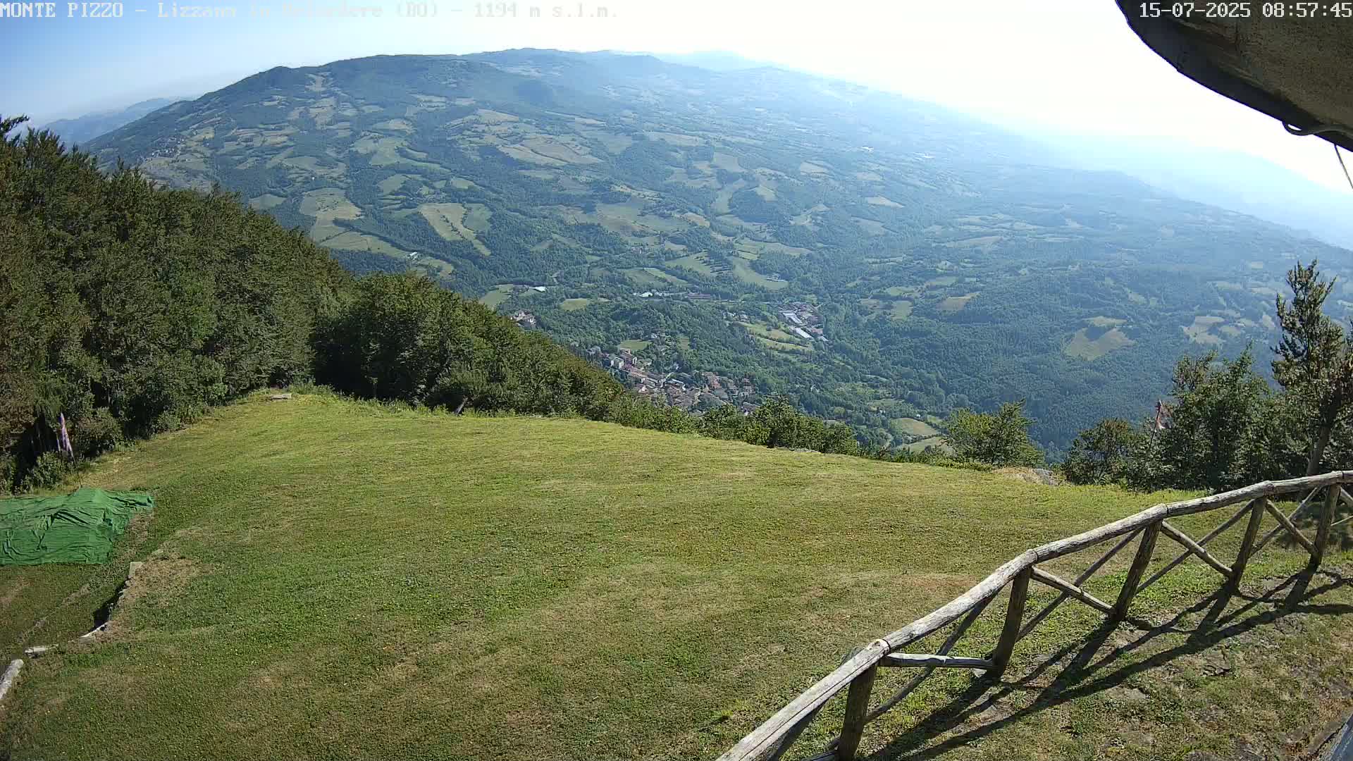 A grassy area overlooks a vast valley of green hills and a small town under a hazy sky.