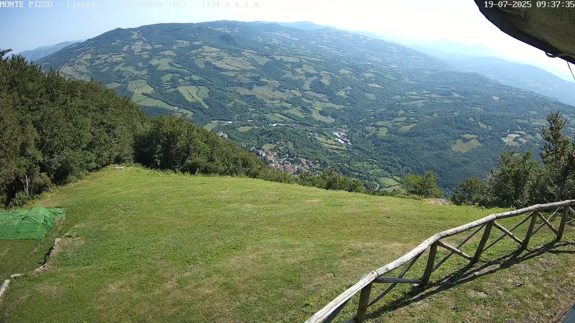 A grassy hilltop with a wooden fence overlooks a vast, verdant valley on a clear, sunny day.