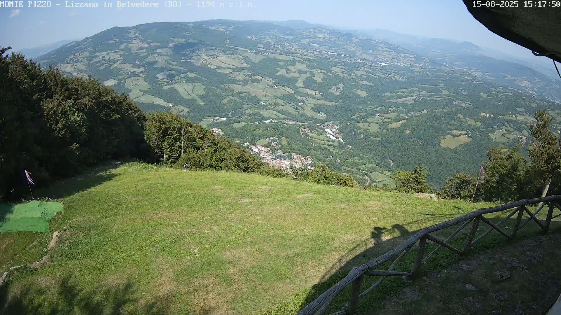 A grassy area with a wooden fence overlooks a valley of green hills and a small town under a clear, sunny sky.
