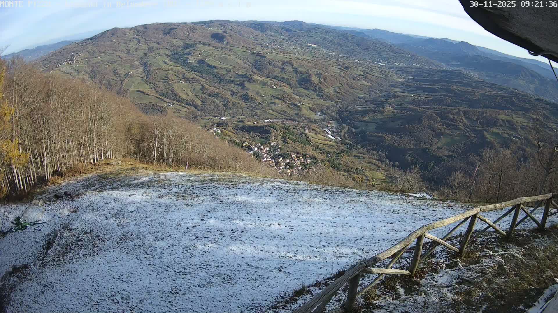 A wide-angle view from a snowy hilltop reveals a vast, rolling landscape with a village nestled in the valley, surrounded by bare trees and distant mountains, all under a clear, sunny sky.