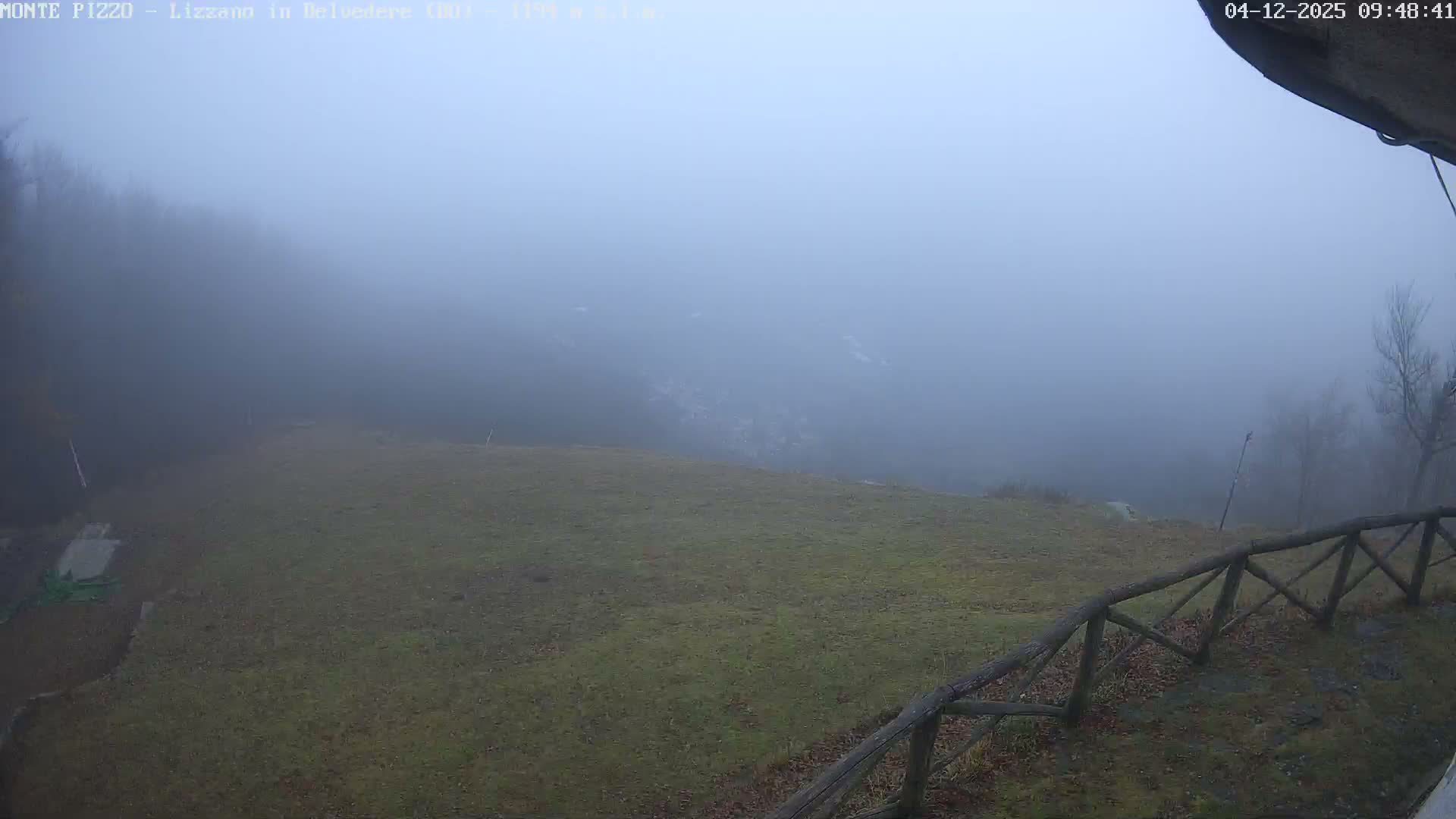 A foggy outdoor landscape features a grassy hillside with a rustic wooden fence in the foreground, while distant bare trees and mountains are heavily obscured by dense mist, resulting in very low visibility.