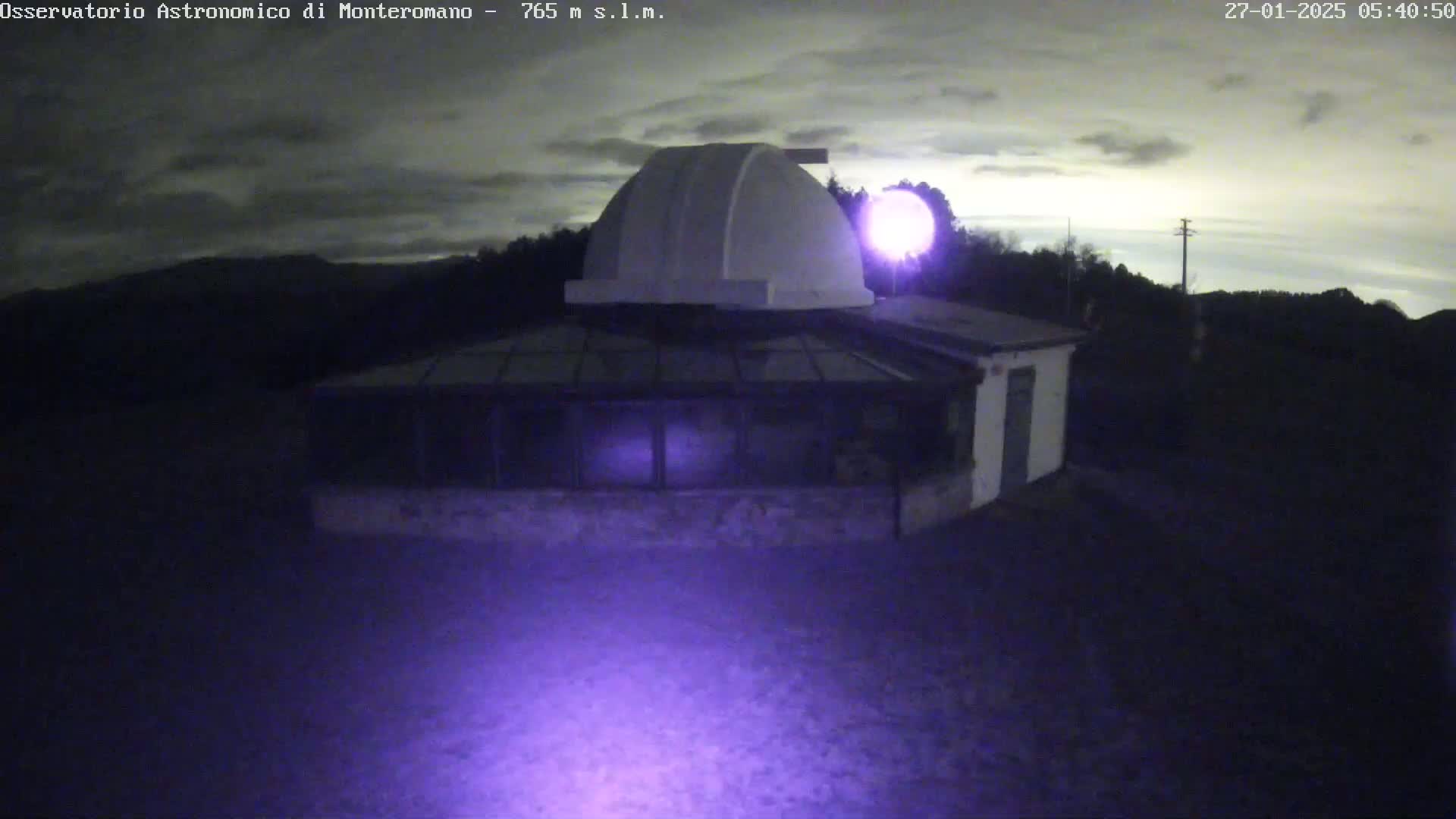 Under a cloudy night sky, a low-lying observatory building with a dome sits on a dark, hilly landscape.