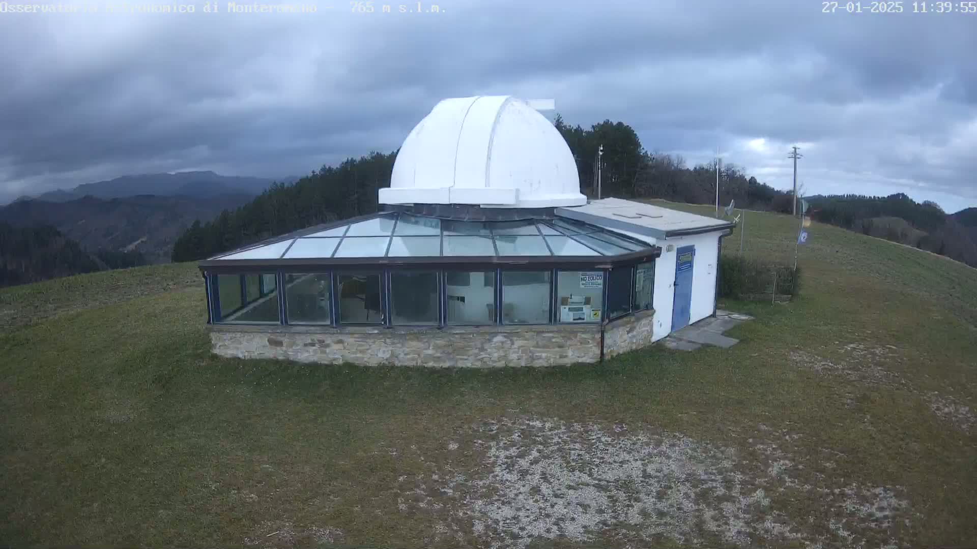 An observatory with a white dome sits atop a grassy hill under a cloudy sky.