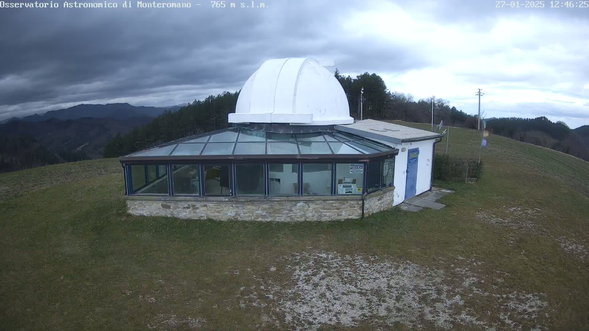 An observatory with a white dome sits atop a grassy hill under a cloudy sky.