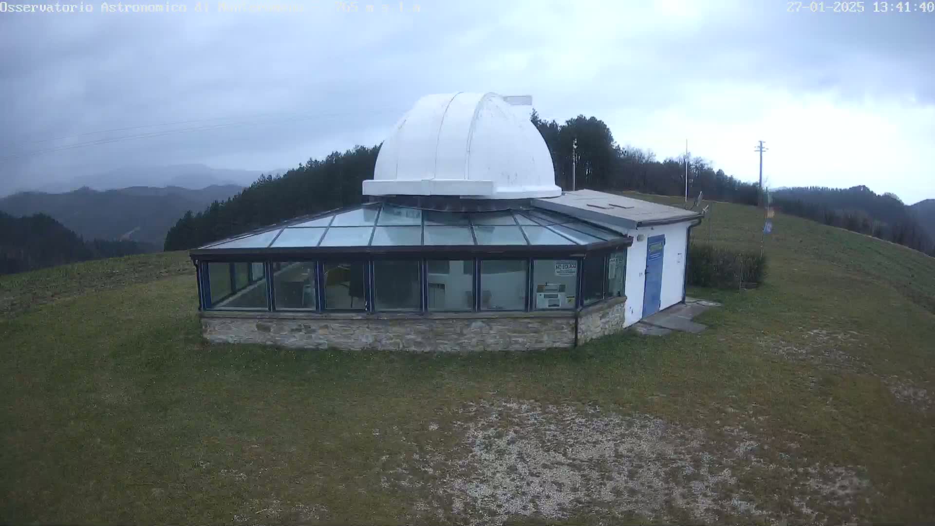 A stone building with a glass sunroom and a white dome on top sits on a grassy hill overlooking a distant, overcast mountain range.