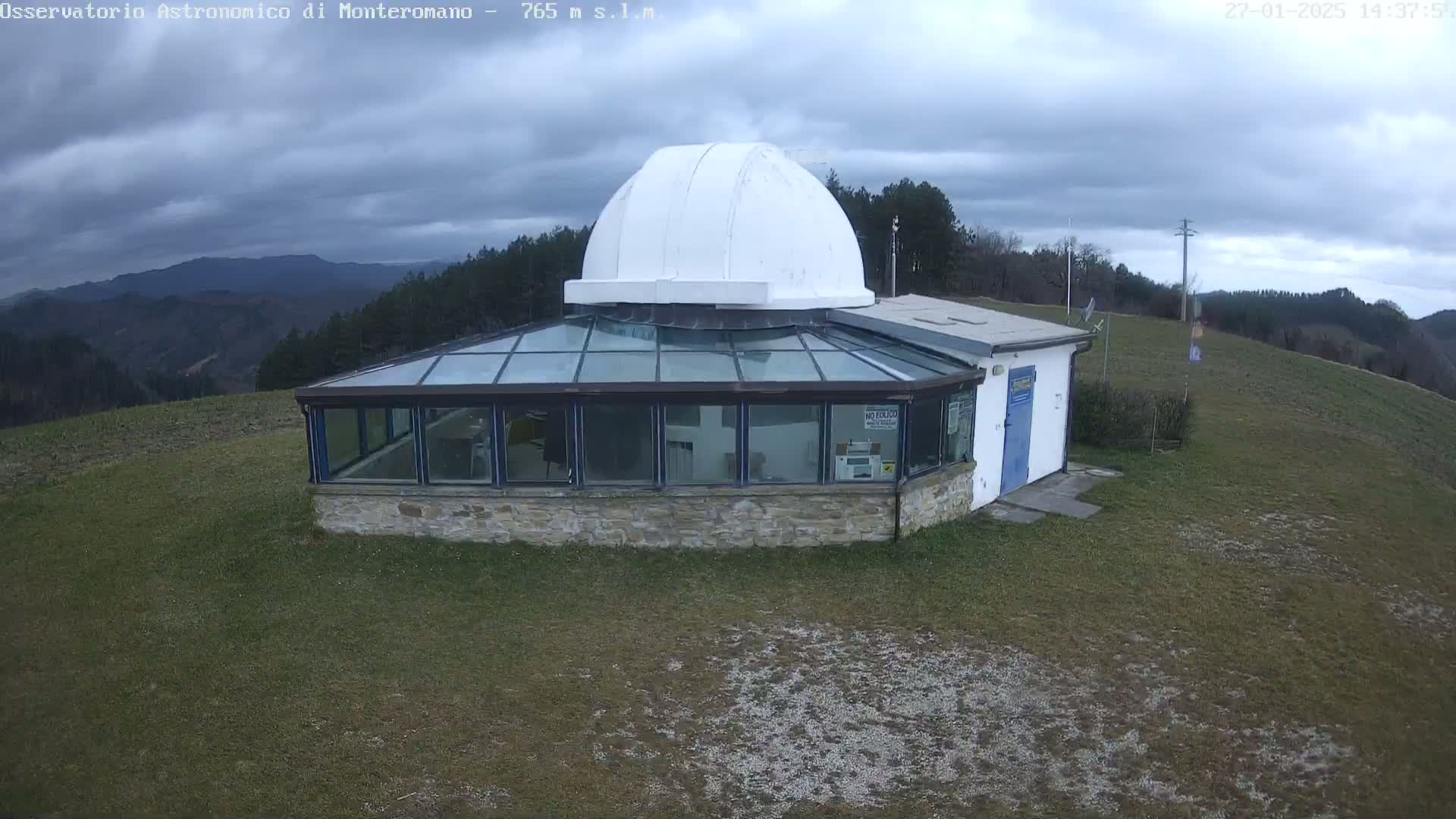 A stone building with a glass sunroom and a white dome on top sits on a grassy hill under a cloudy sky.