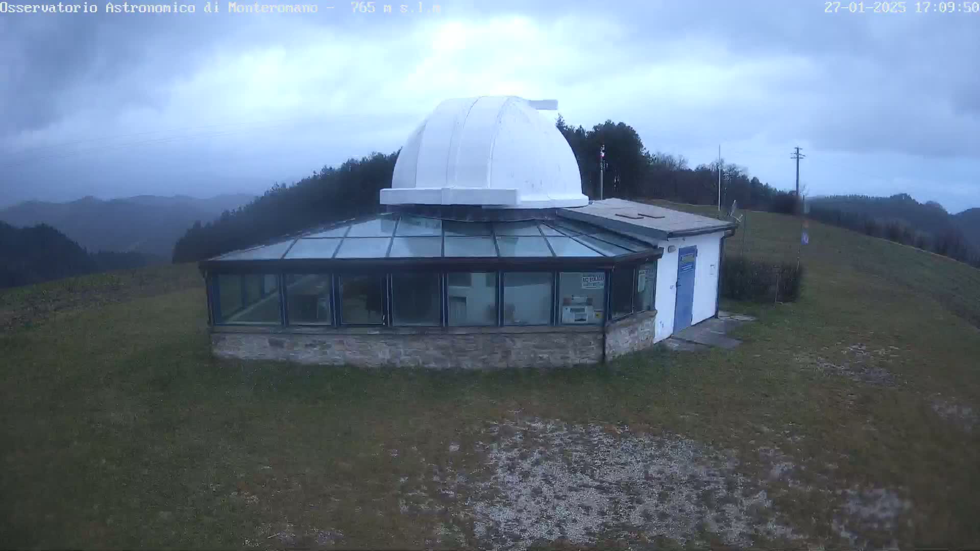 A white domed observatory building with a glass-roofed addition sits on a grassy hill under an overcast sky.