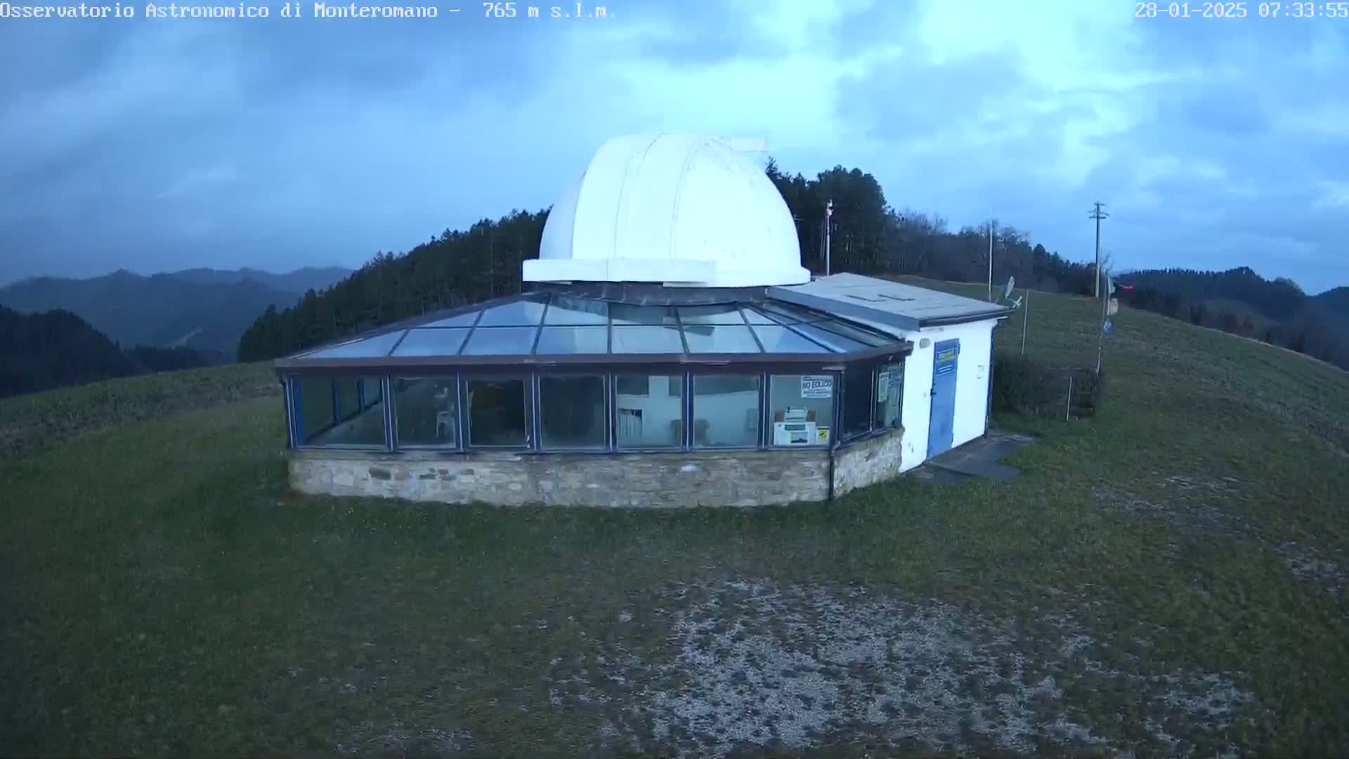 An observatory with a glass-enclosed viewing area and a white dome sits atop a hill under a cloudy sky.