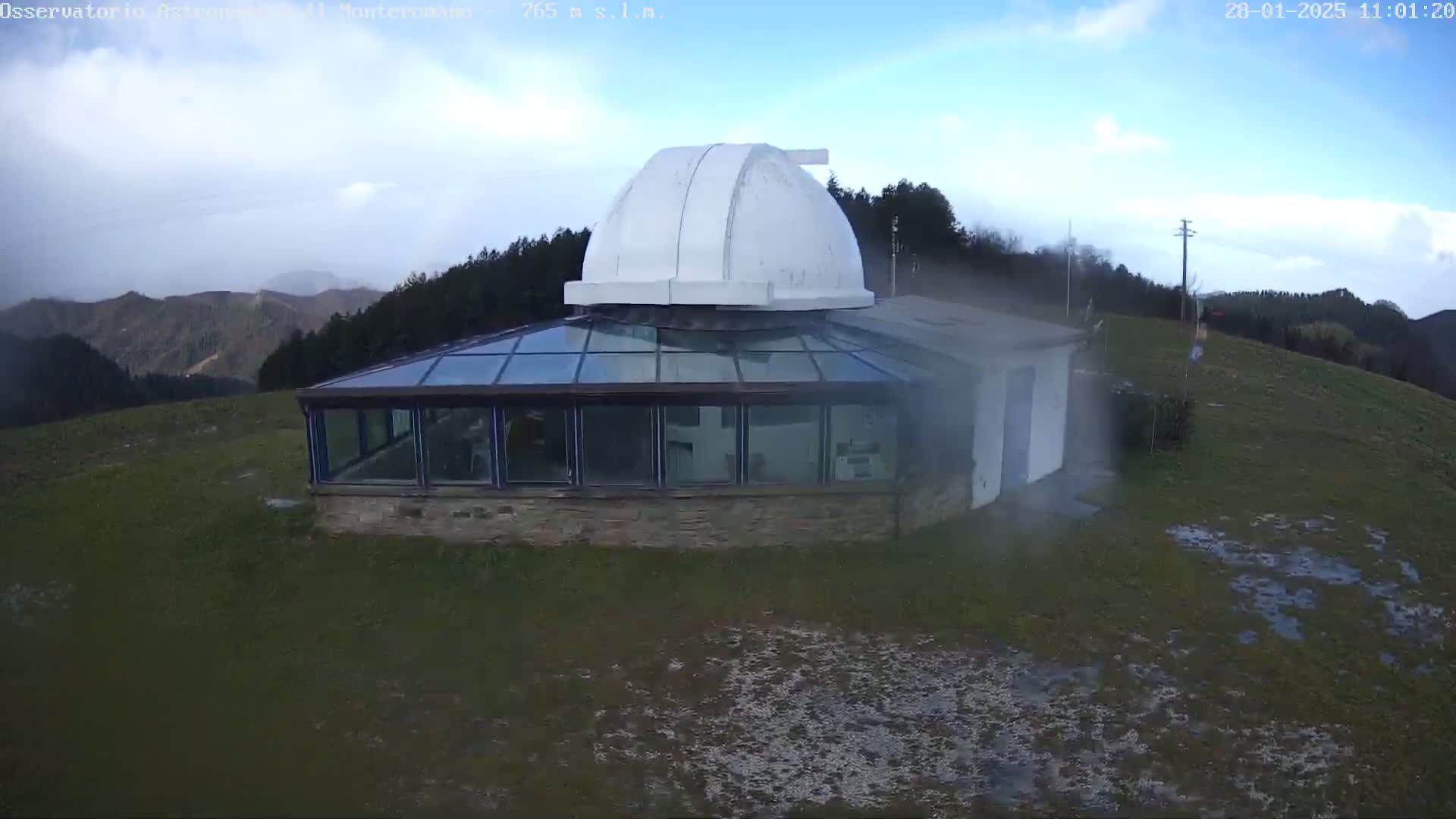 An observatory with a glass-roofed addition sits on a grassy hill under a partly cloudy sky.