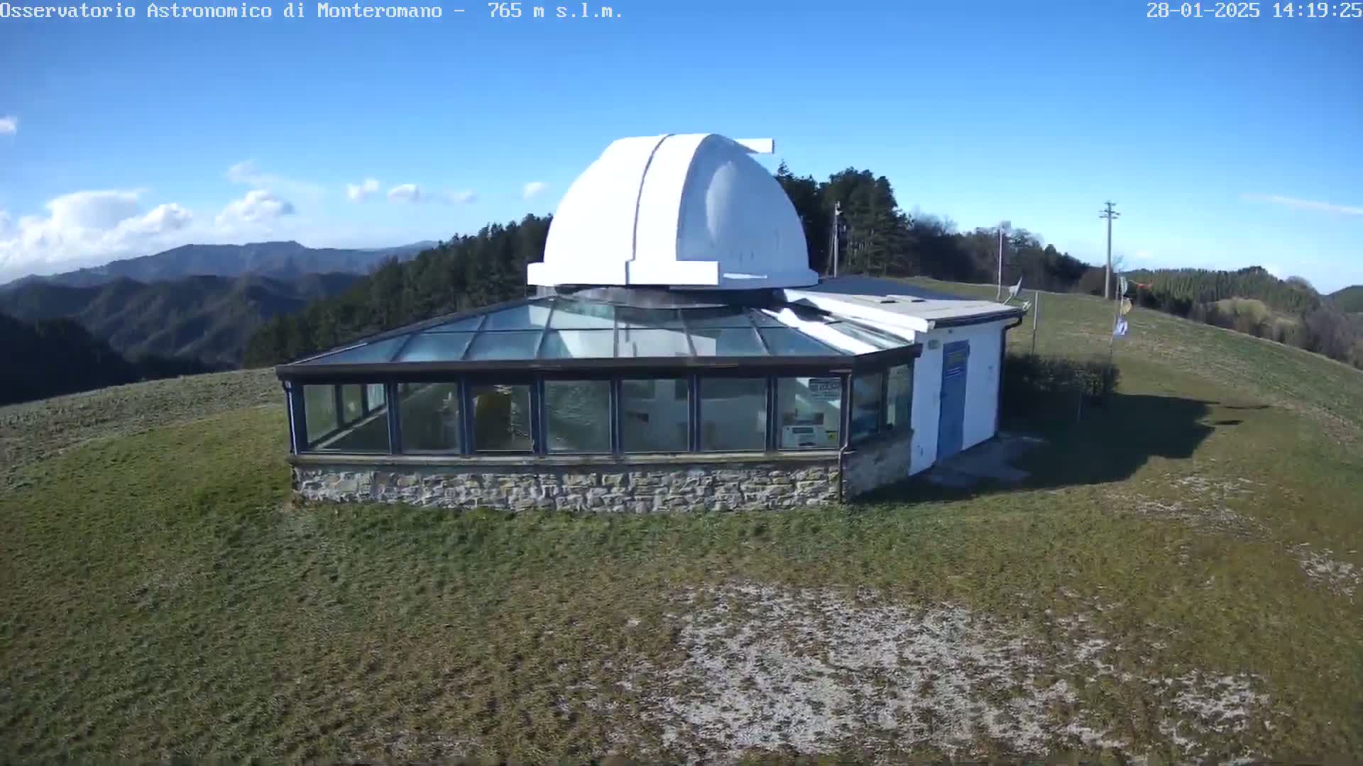 A white observatory dome sits atop a stone building with a glass roof, overlooking a grassy hillside under a mostly clear blue sky.