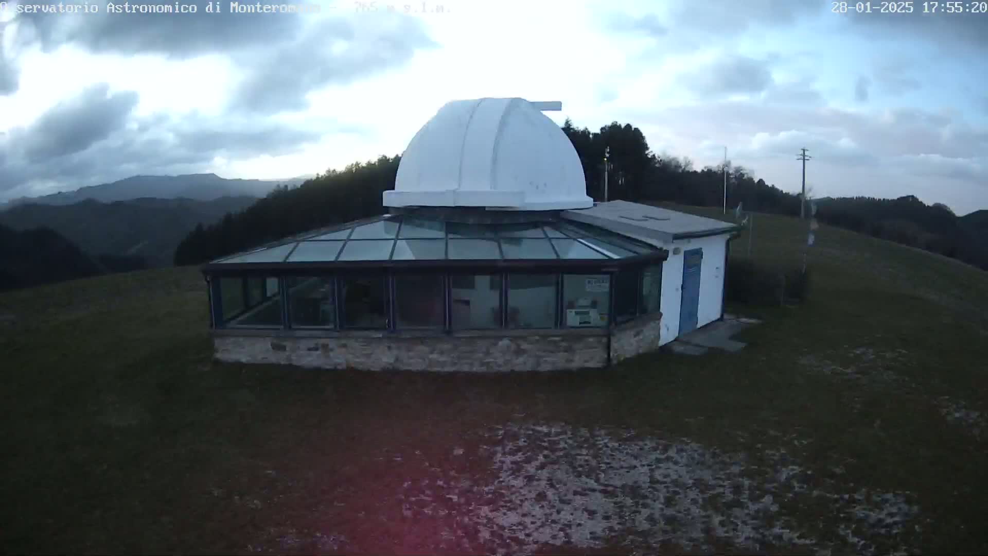 An observatory with a white dome sits atop a hill under a cloudy sky, overlooking a range of dark green mountains.