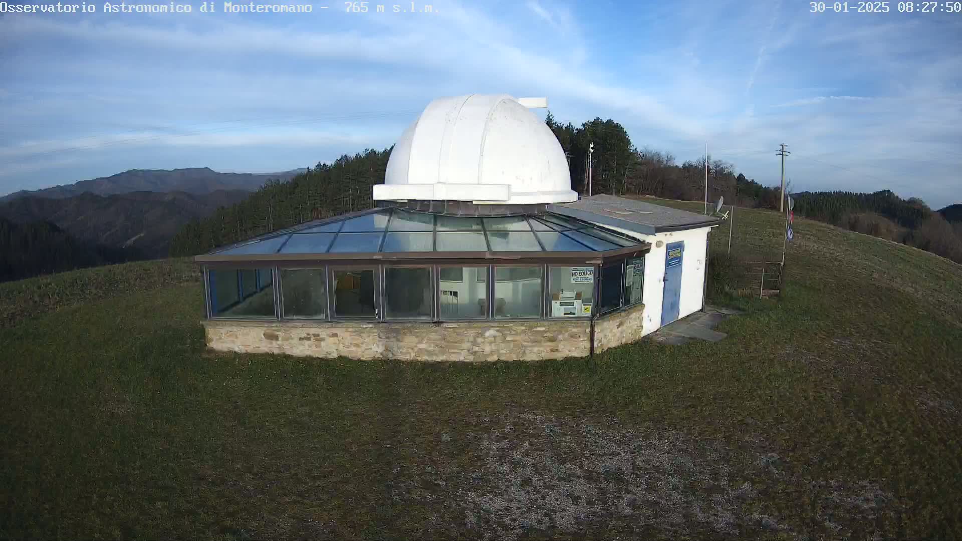 A stone building with a glass roof and a white dome on top sits on a grassy hill under a mostly clear blue sky.