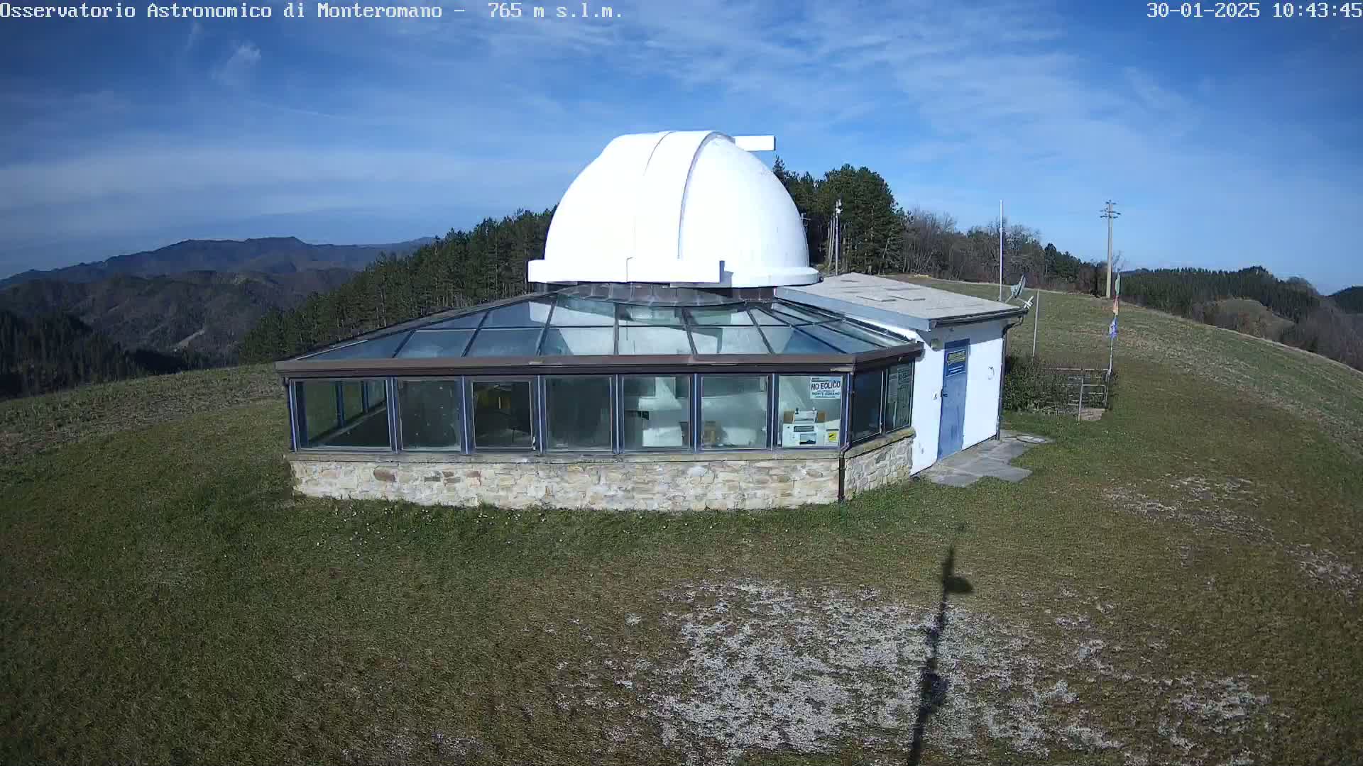 A white observatory dome sits atop a stone building with a glass enclosure, overlooking a grassy hillside under a mostly clear blue sky.