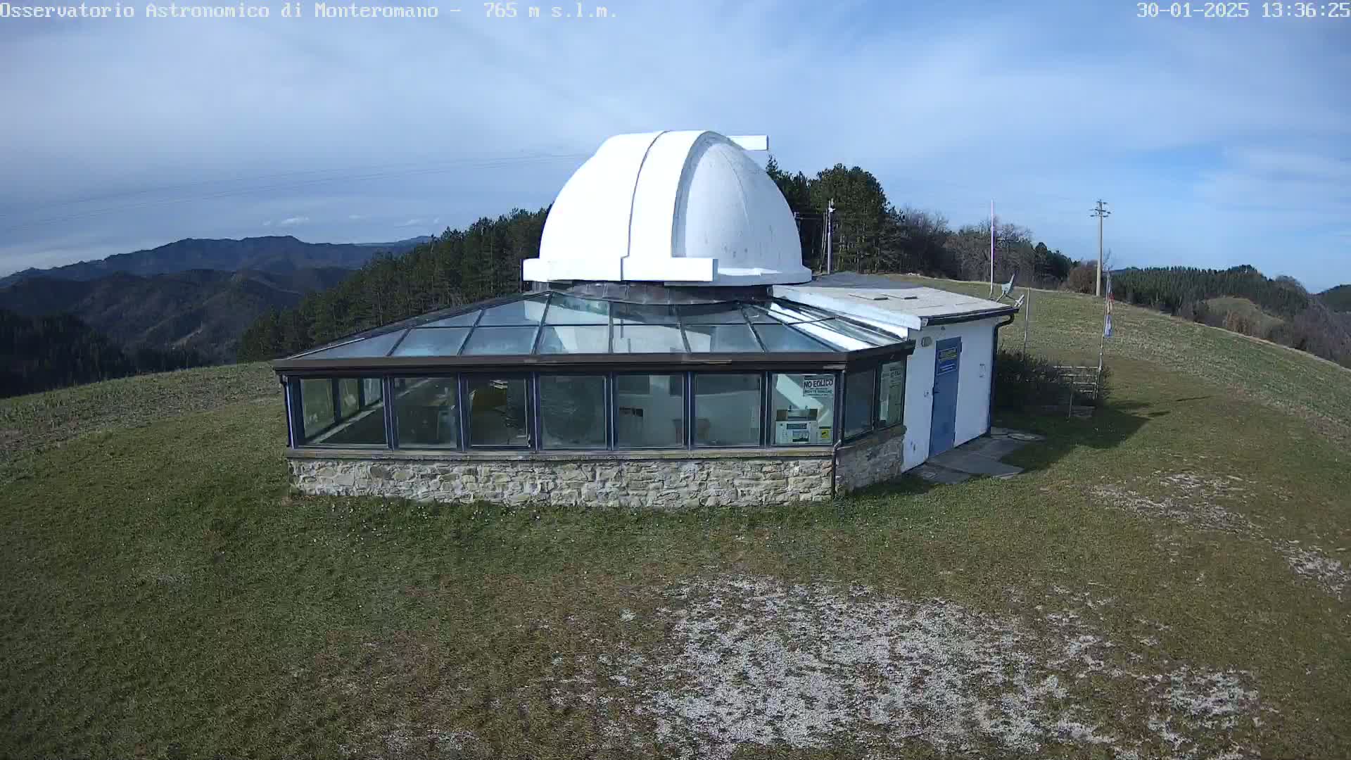 An astronomical observatory with a white dome sits atop a grassy hill under a mostly clear, sunny sky, overlooking a mountainous landscape.