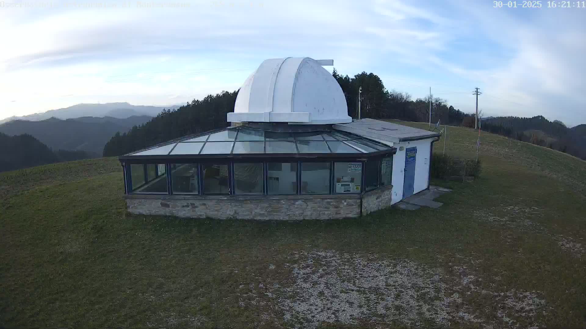 A stone building with a glass conservatory and a white dome sits atop a grassy hill overlooking a range of mountains under a partly cloudy sky.