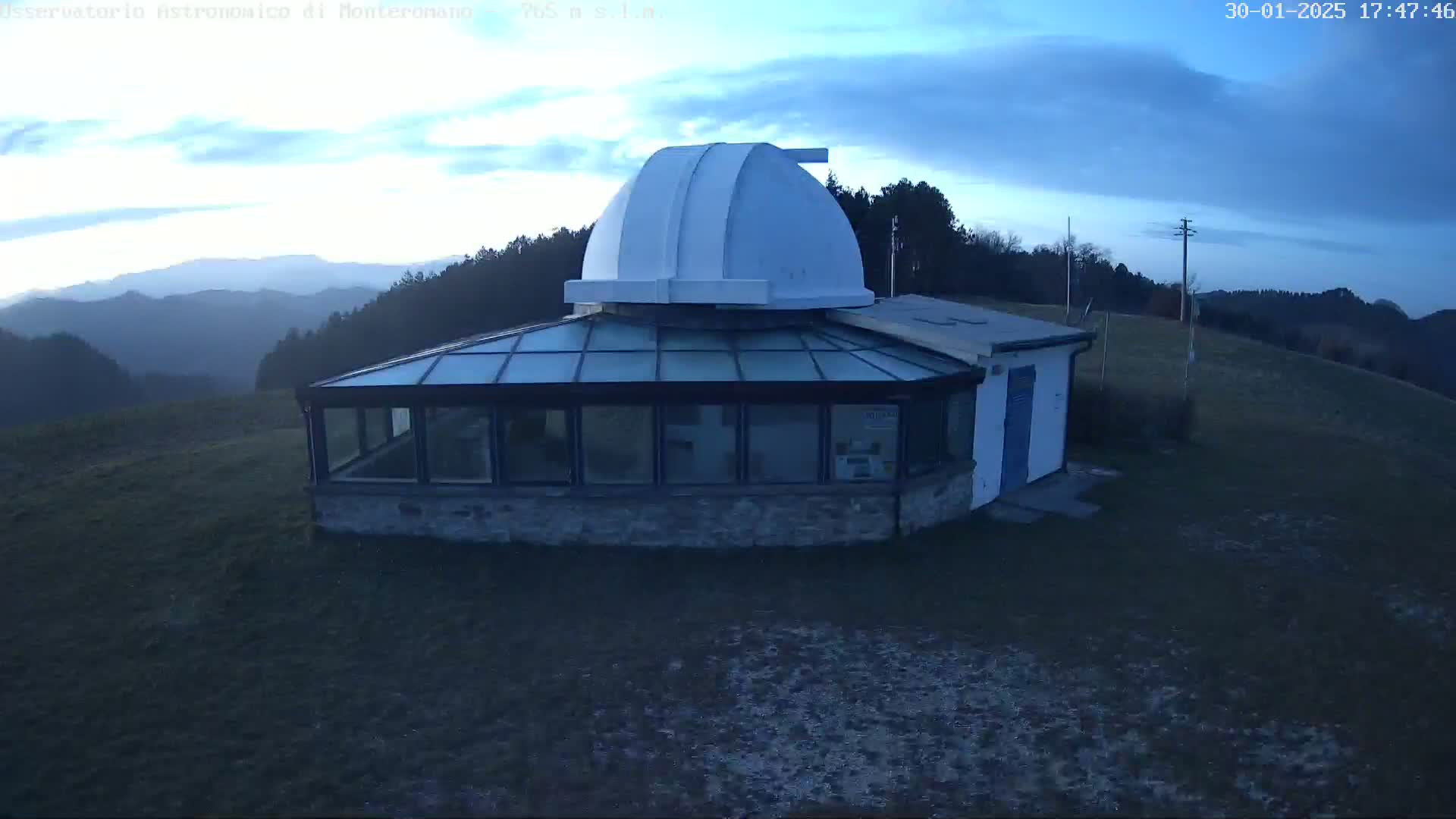 An observatory with a white dome sits atop a grassy hill overlooking a mountain range under a mostly cloudy sky.