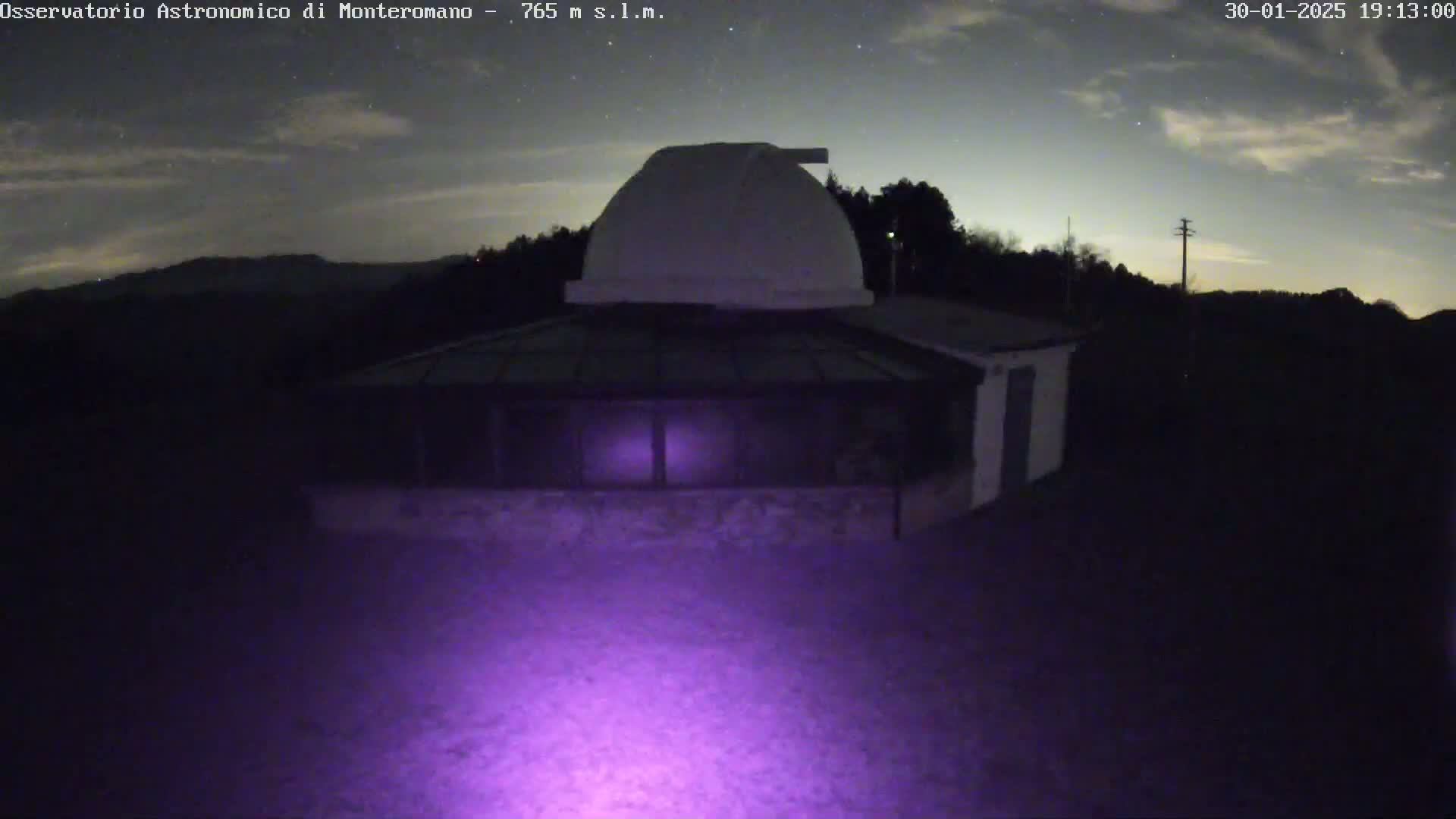 A low-light nighttime image shows an observatory building with a dome under a partly cloudy sky.