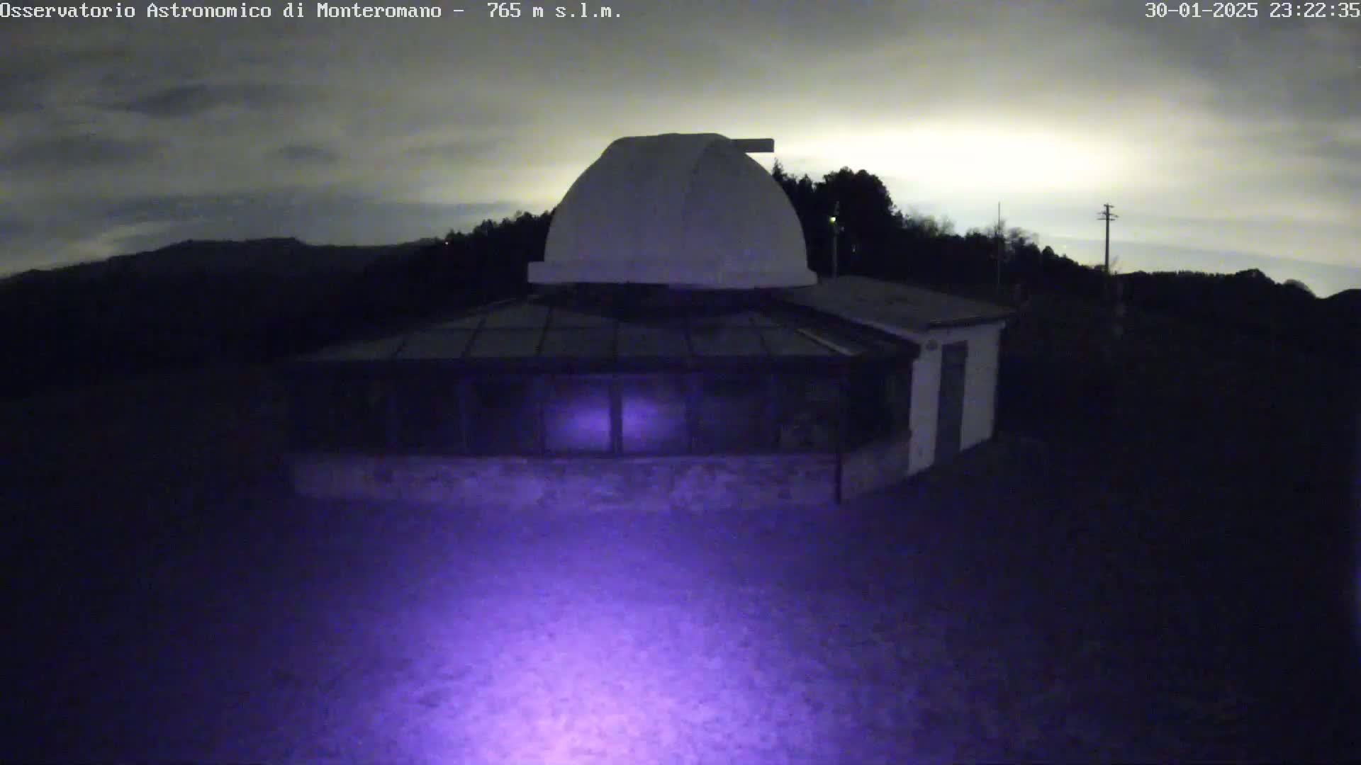 A low-light nighttime image shows a building with a dome on top, surrounded by dark trees under a cloudy sky.