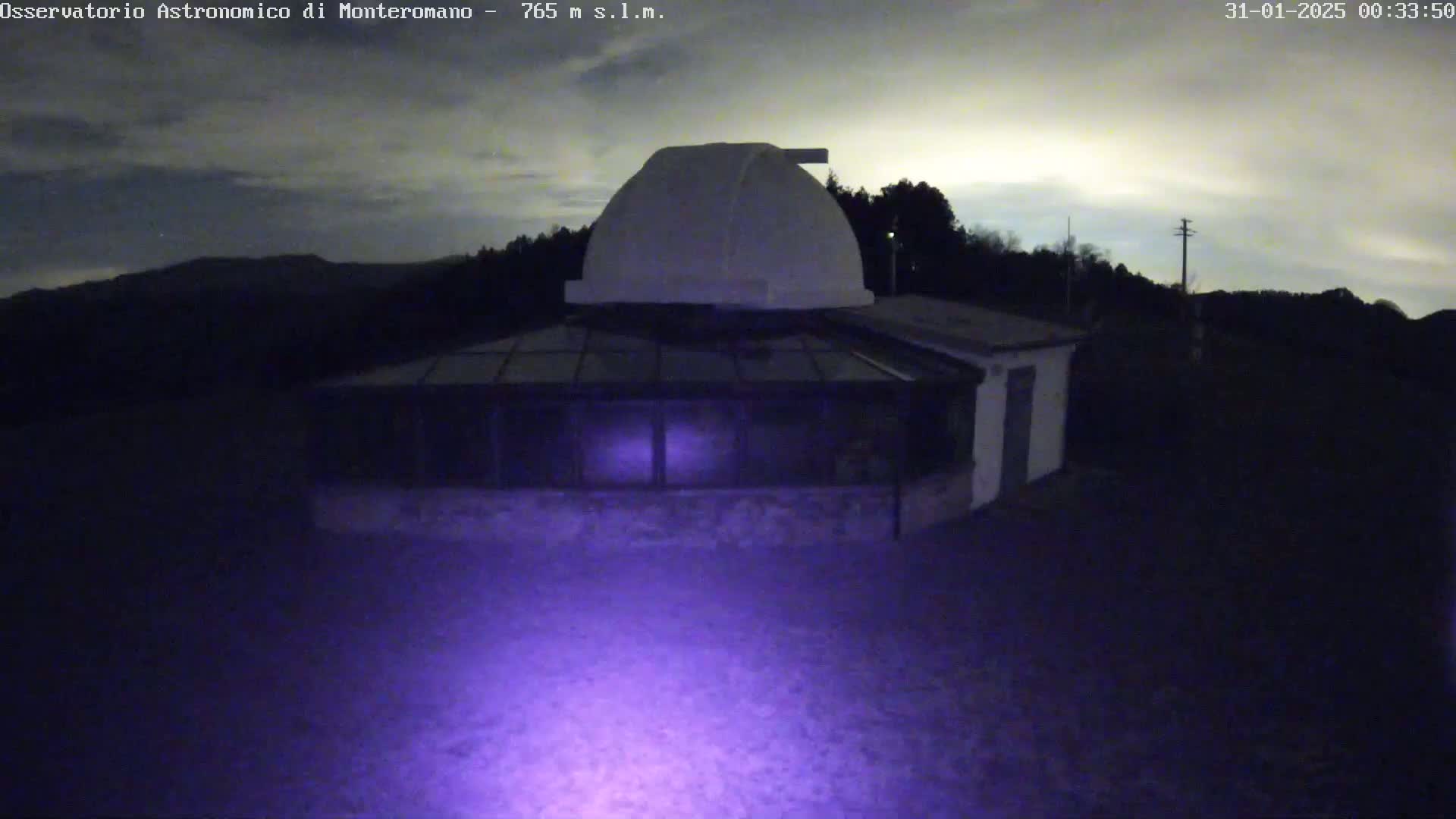 A low-light nighttime image shows a white dome-shaped observatory building with a glass roof section, situated on a hill under a mostly clear, dark sky.