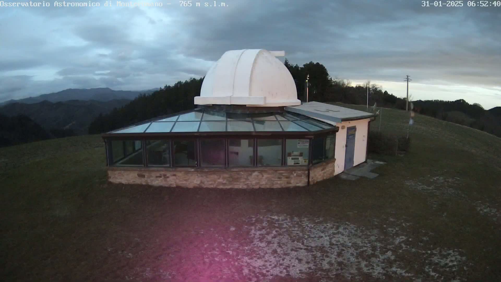 A white-domed observatory building with a glass sunroom sits on a hilltop overlooking a range of mountains under a cloudy sky.