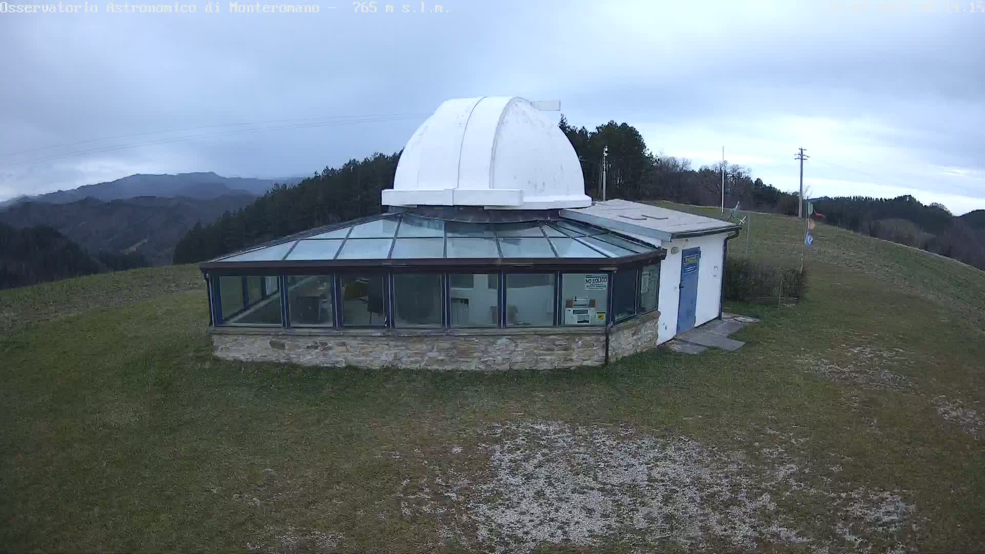 A white-domed observatory building with a glass-roofed addition sits on a grassy hill overlooking a distant mountain range under an overcast sky.