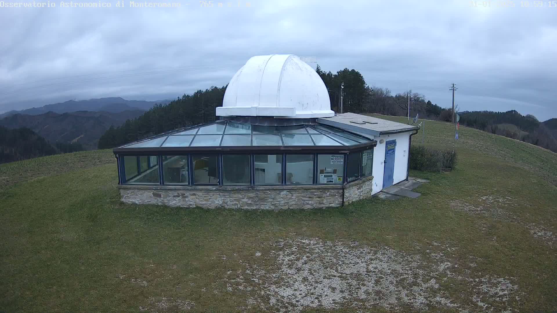 A stone building with a glass sunroom and a white dome sits on a grassy hill overlooking a range of mountains under a cloudy sky.