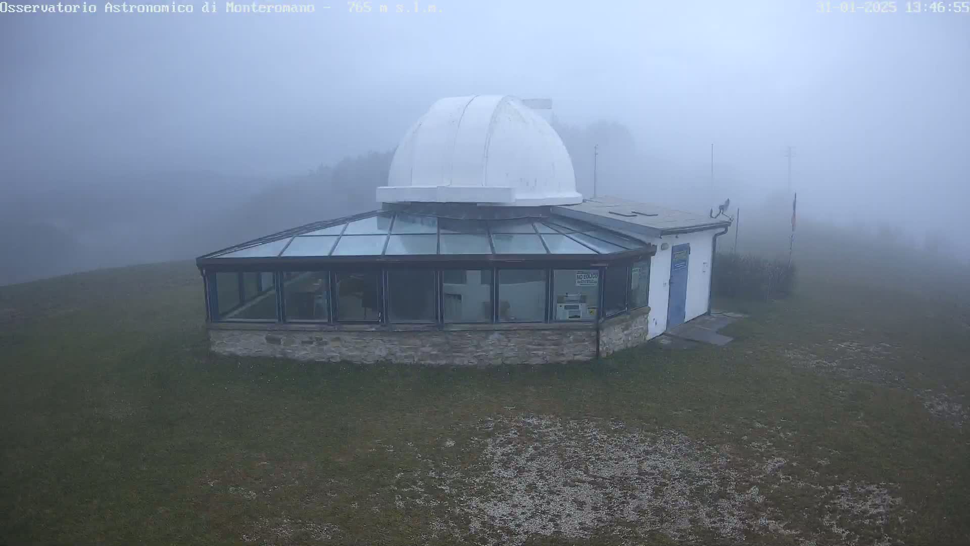 A white-domed observatory building with a glass-roofed addition sits on a grassy area shrouded in thick fog.