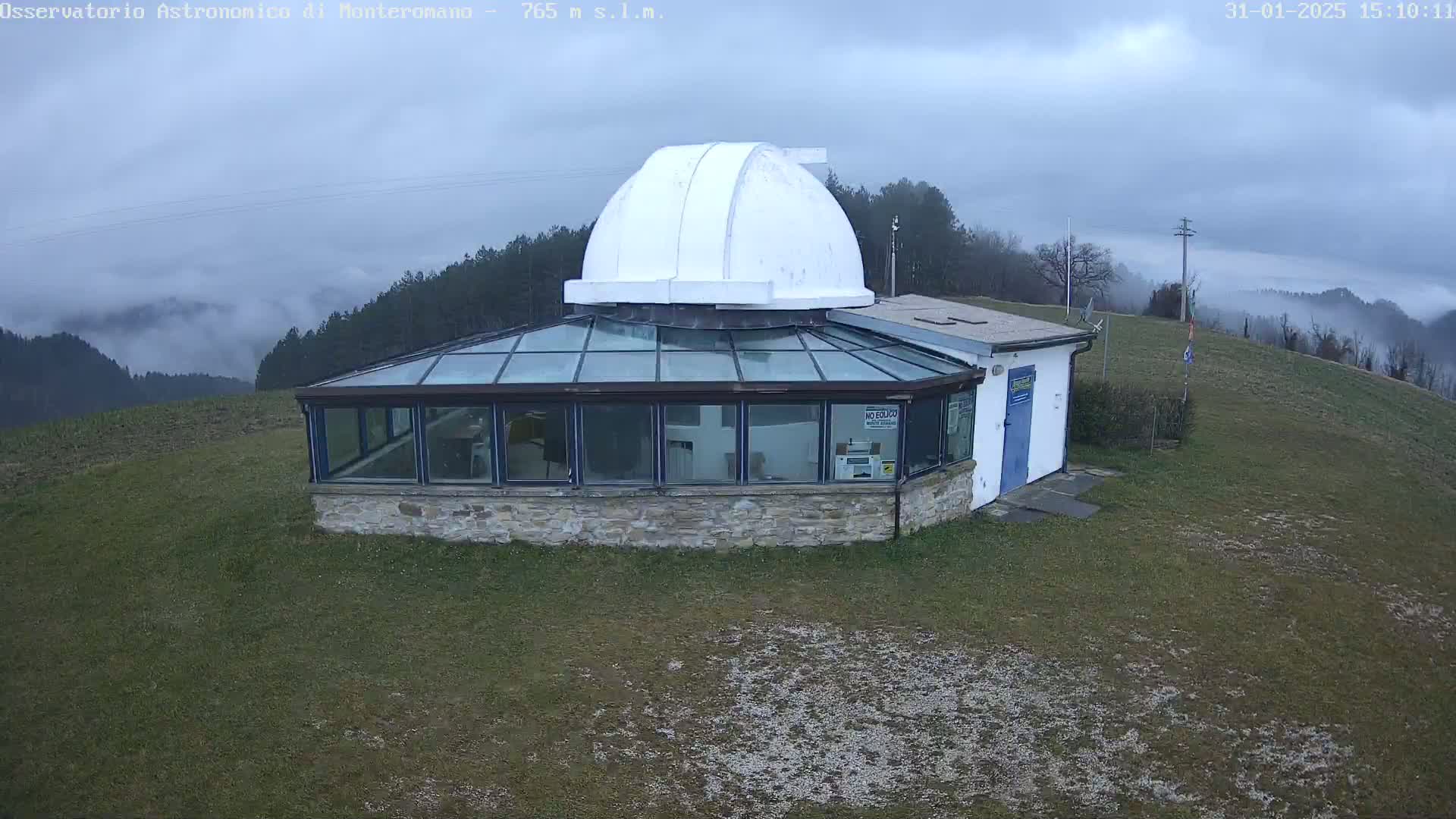 An observatory with a white dome sits on a grassy hilltop under a cloudy sky, with low-lying fog in the distance.
