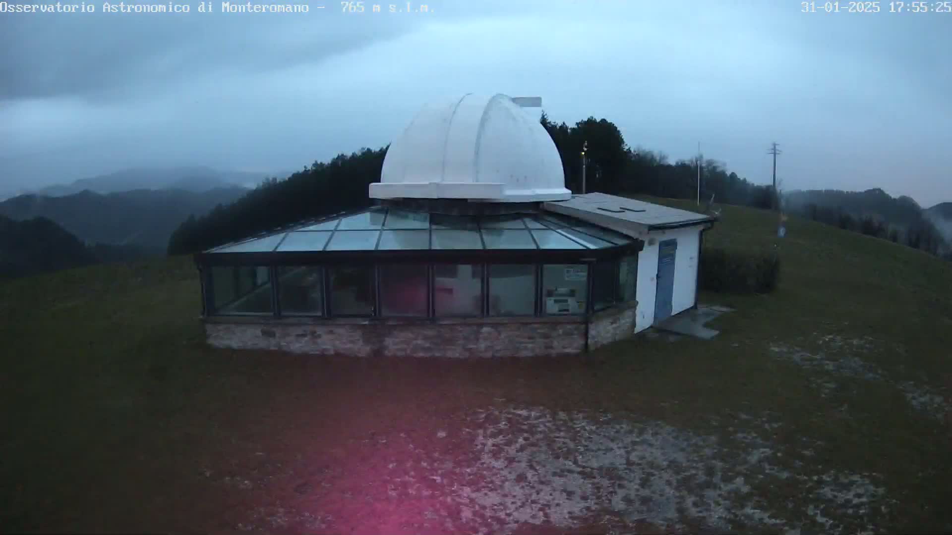 A white domed observatory building with a glass-roofed addition sits on a grassy hill overlooking a hazy mountain range under an overcast sky.