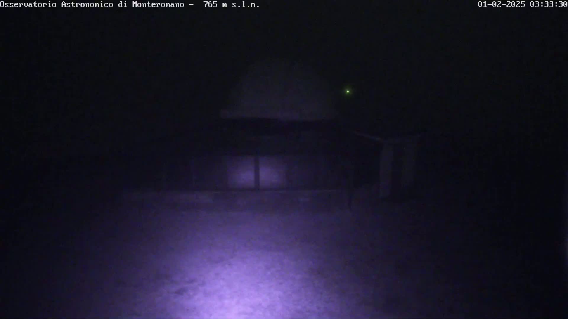 A low-light nighttime image shows a building with a domed roof, illuminated by a faint light source in the foreground, under foggy or hazy conditions.