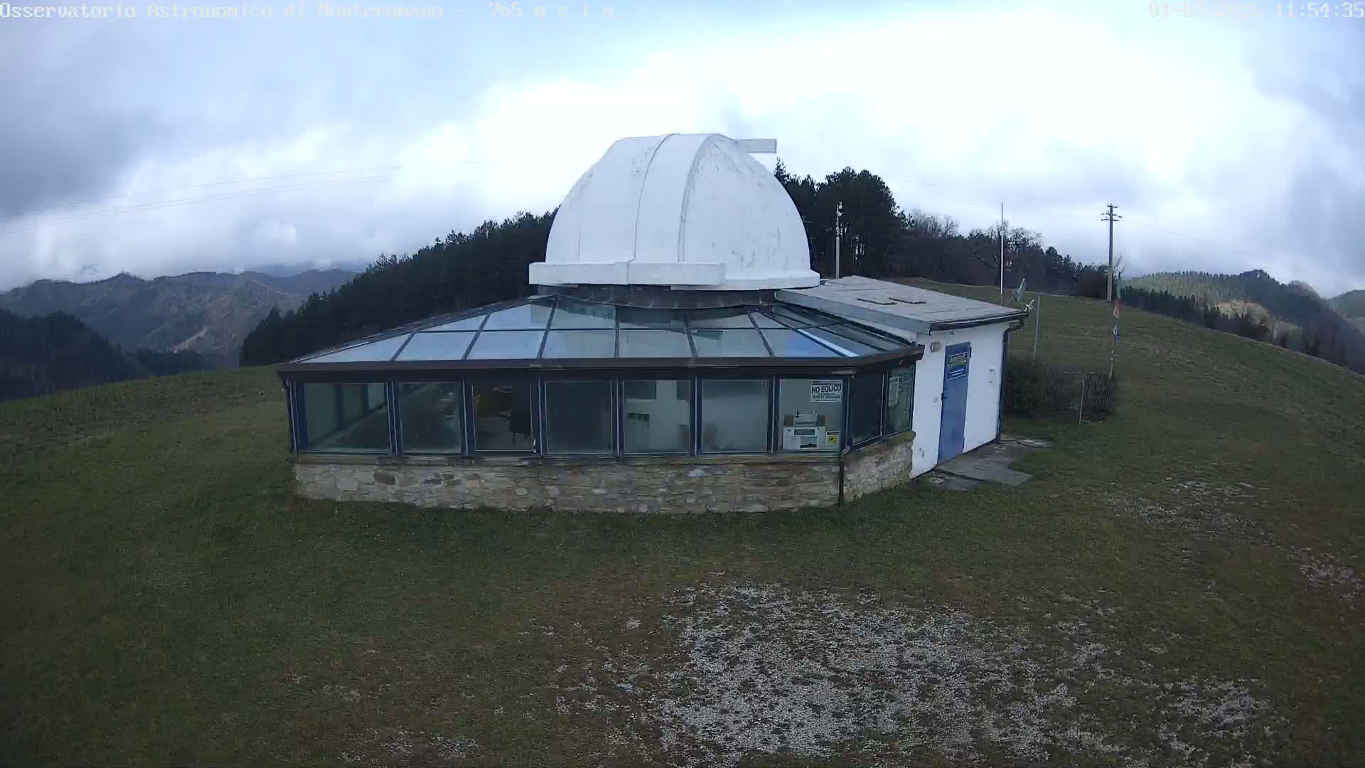 A stone building with a glass-roofed addition and a white dome sits on a grassy hilltop overlooking a partly cloudy sky and distant mountains.