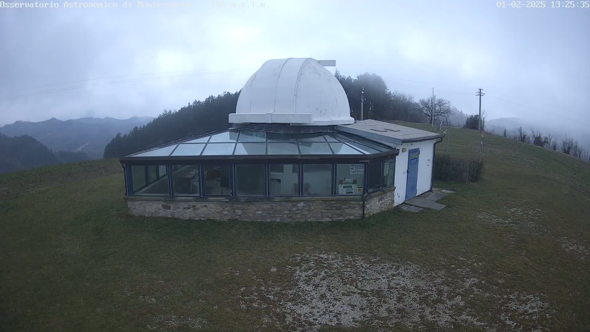 An observatory with a glass-roofed addition sits atop a grassy hill overlooking a foggy mountain range.