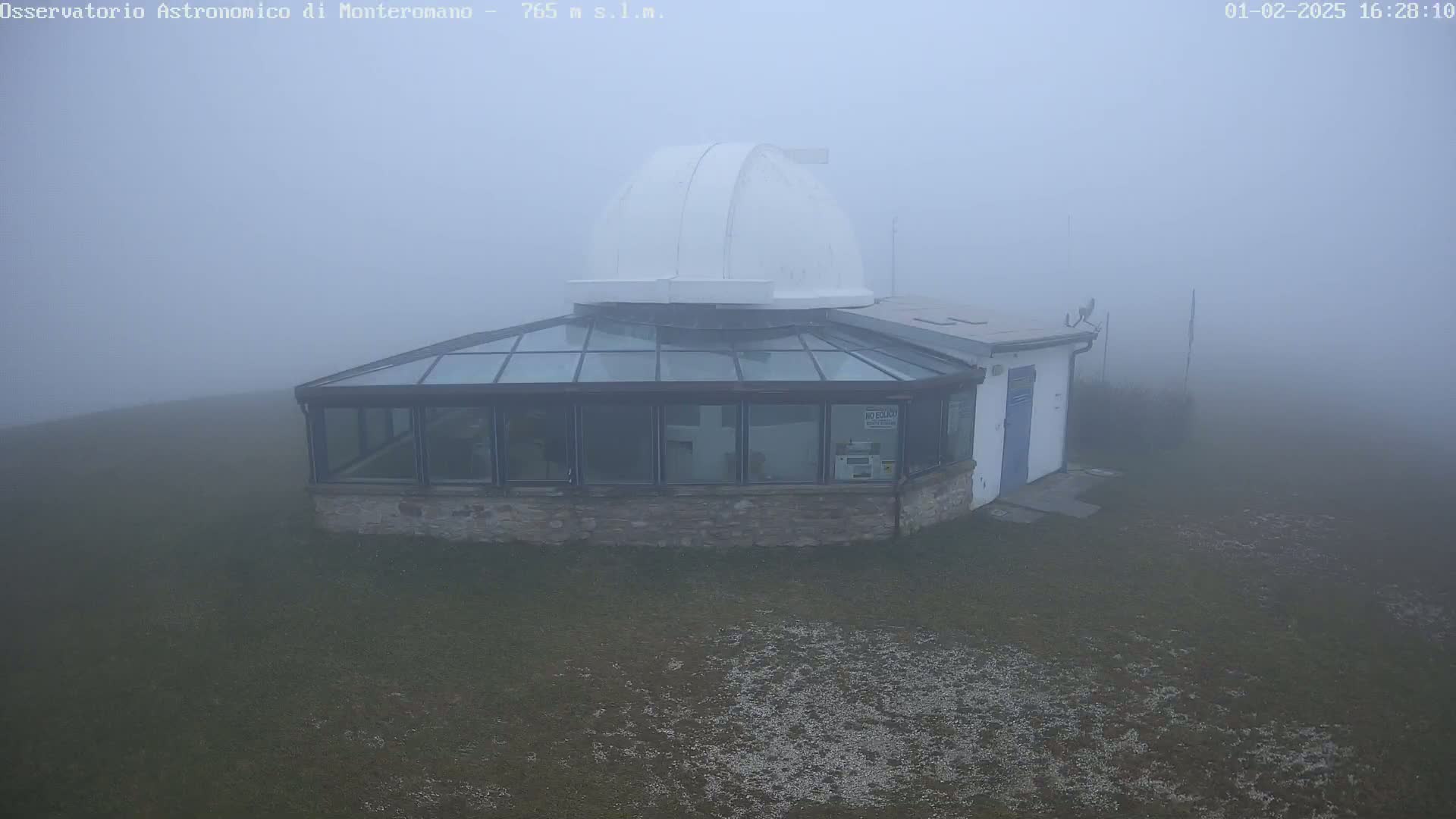 A white dome observatory building with a glass-roofed addition sits on a grassy area in heavy fog.