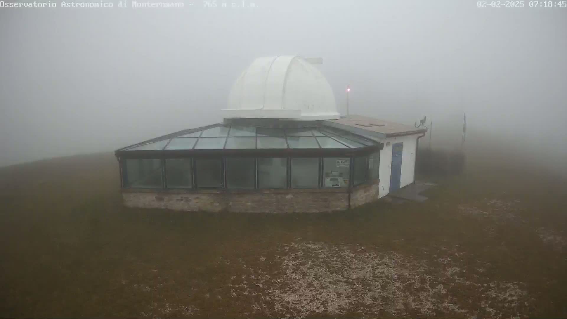 A white-domed observatory building with a glass-roofed addition sits shrouded in dense fog on a flat, brown expanse of land.