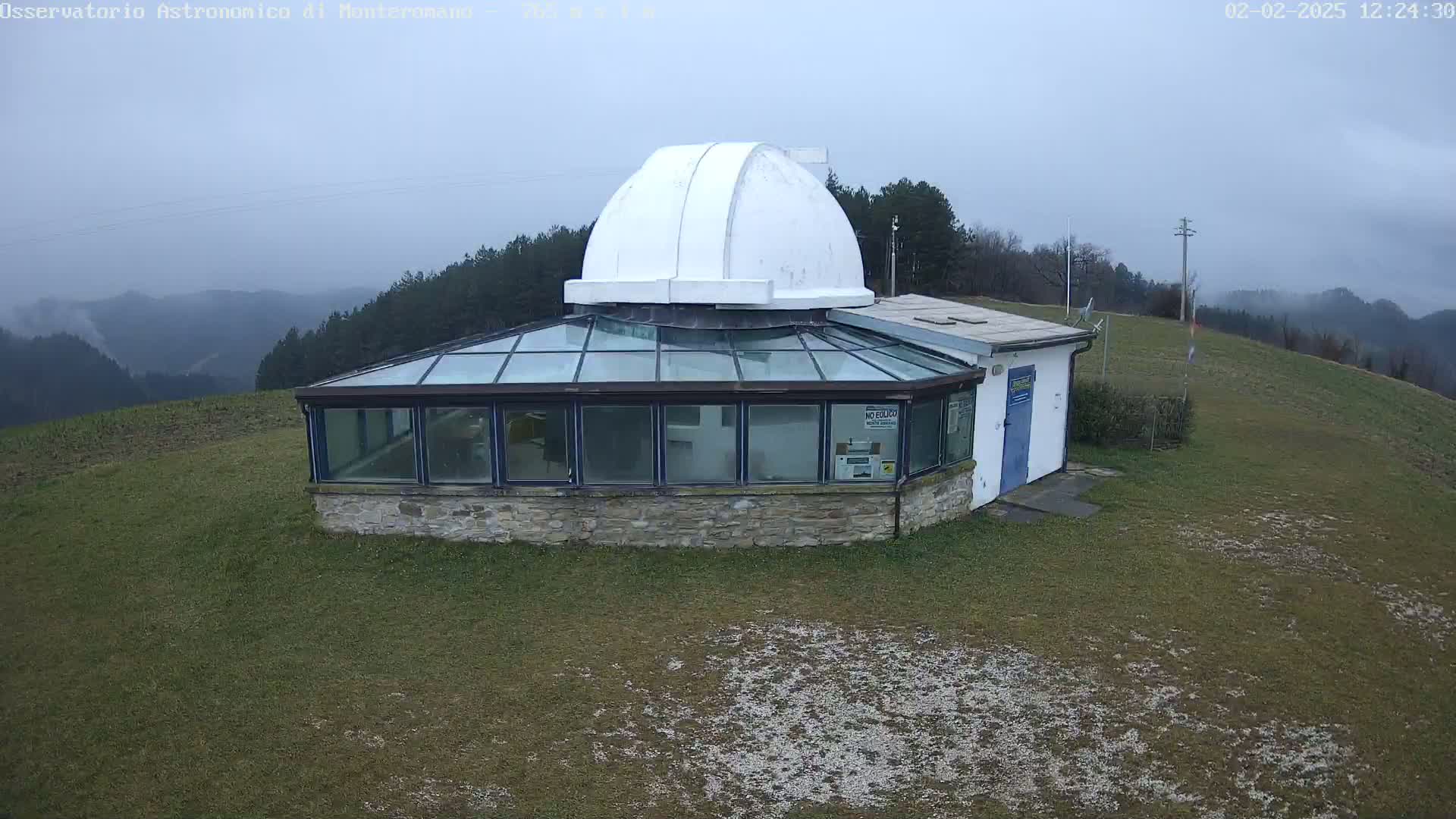 A white domed observatory building with a glass-roofed addition sits on a grassy hill overlooking hazy, distant mountains under an overcast sky.